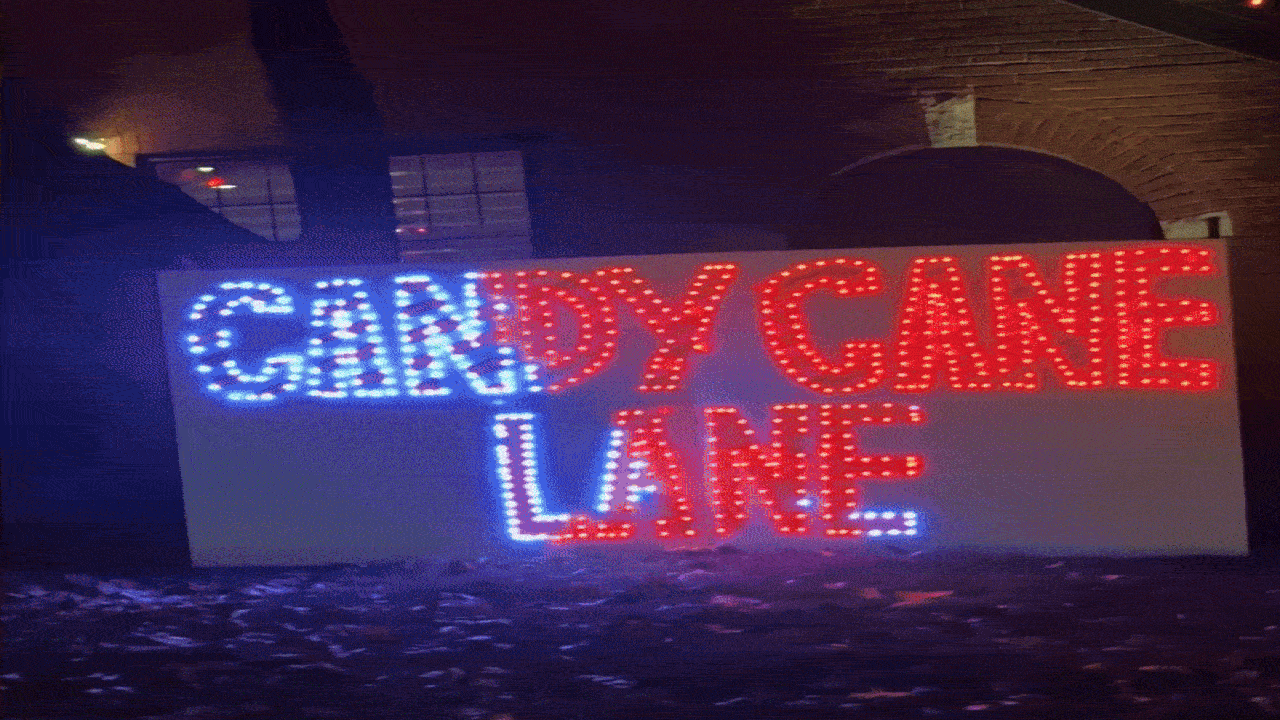 A light-up sign at night reading "CANDY CANE LANE" with blue and red lights, set against a dark background with a brick arch nearby.