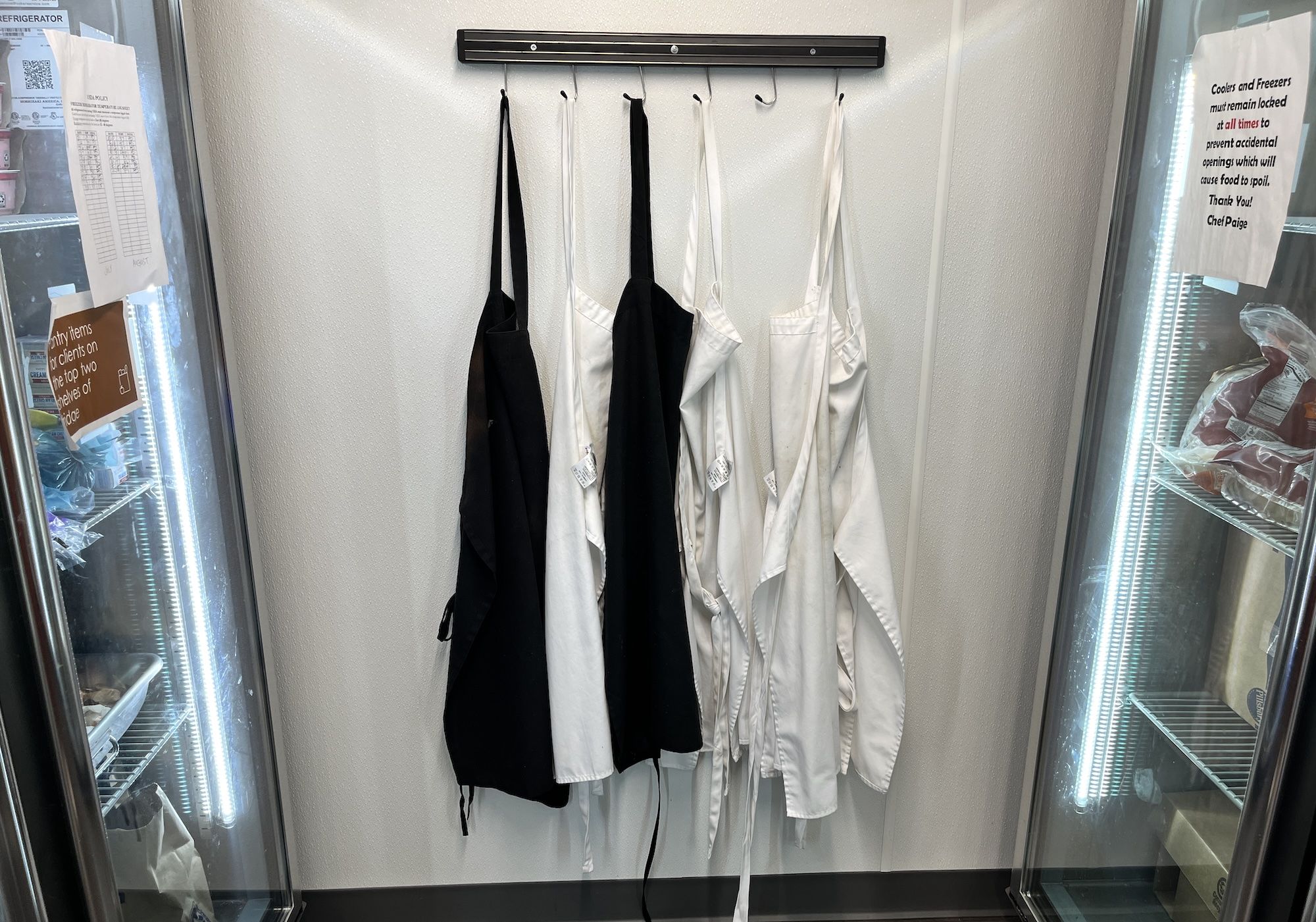 Inside a small walk-in cooler, white chef jackets and black aprons hang from a metal rod with hooks. Glass doors show posted signs and a list inside.