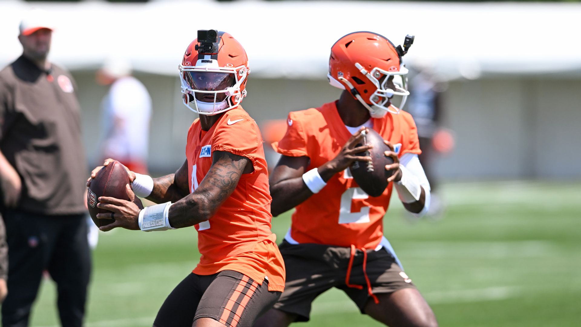 Two football players in orange jerseys and white helmets practice on a sunny field. The foreground player grips a football with a GoPro mounted on his helmet; a teammate behind him also holds a ball.