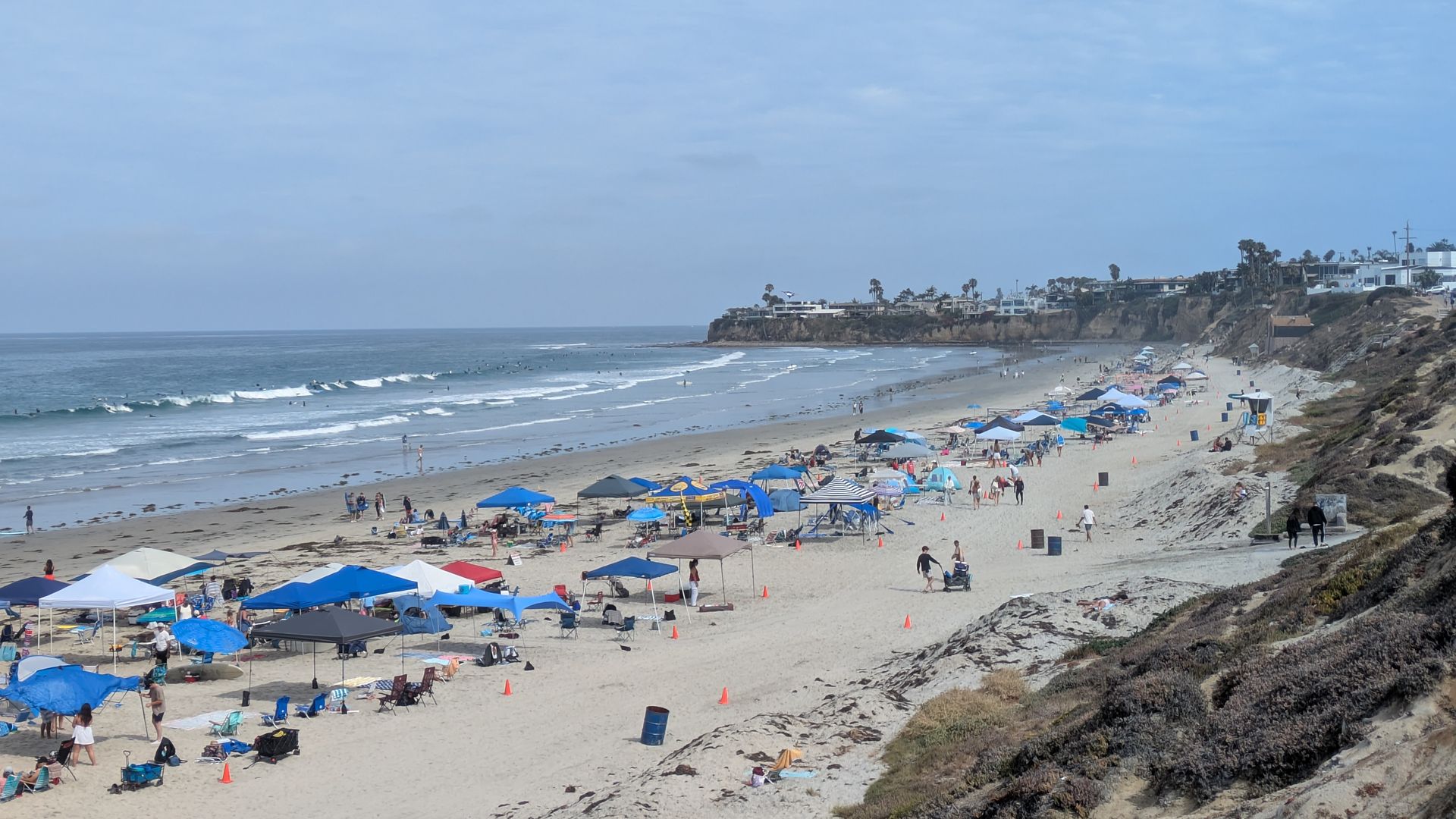 Sunny beach scene with numerous colorful umbrellas and people along the sandy shore, gentle waves in the ocean to the left, and a hillside with houses and palm trees to the right under a blue sky.