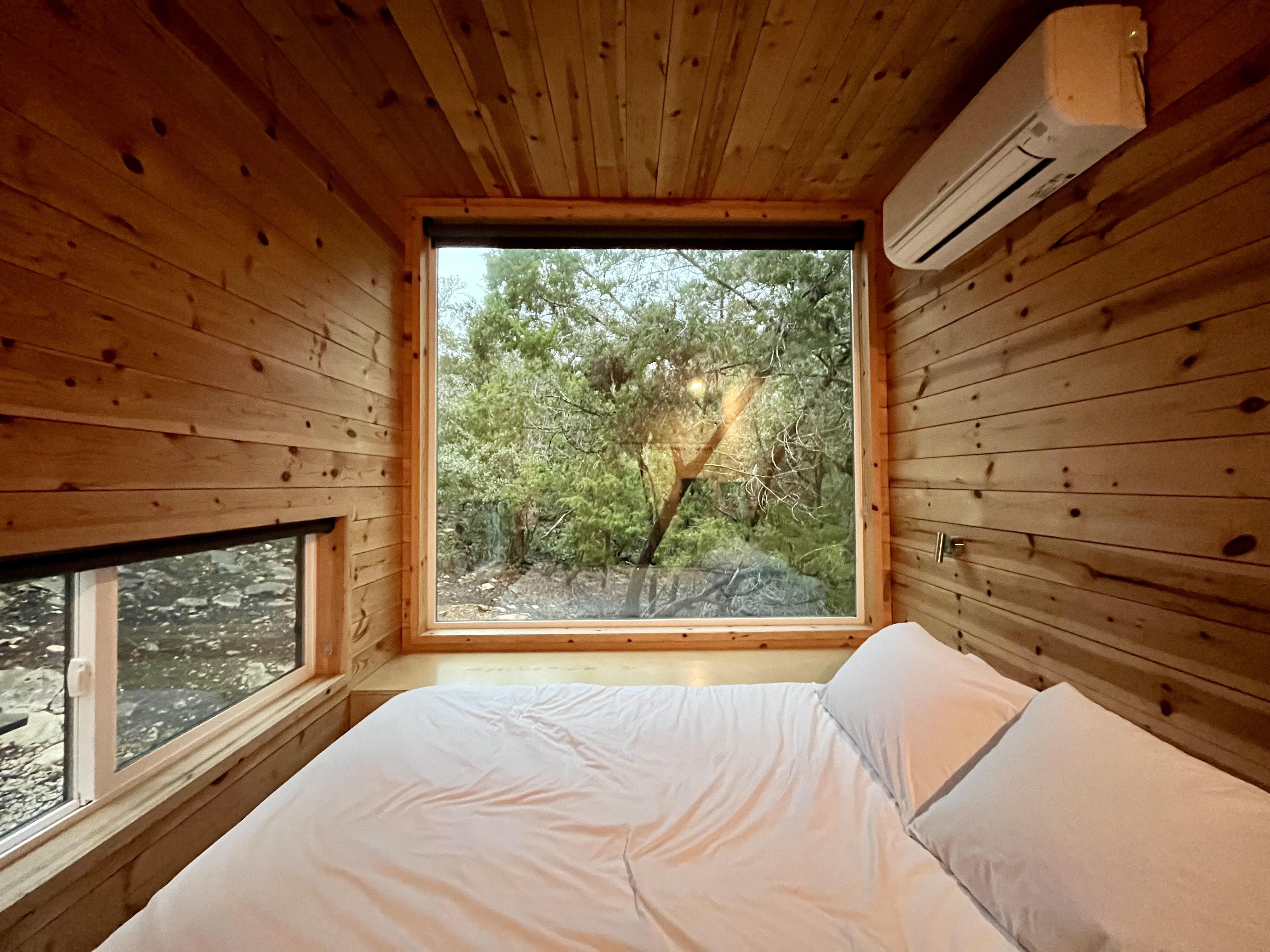 A pine-walled tiny cabin with a queen bed with white bedding has a full-length window that shows a forest outside.