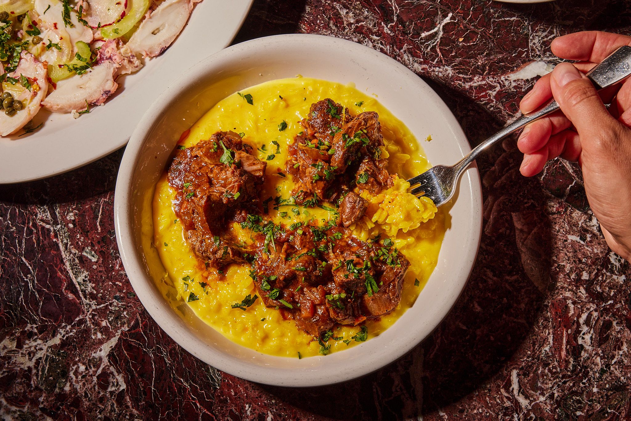 Hand holding a fork with yellow risotto and braised meat in a white bowl, garnished with green herbs, on a dark red marble surface. A plate with sliced food is partially visible.
