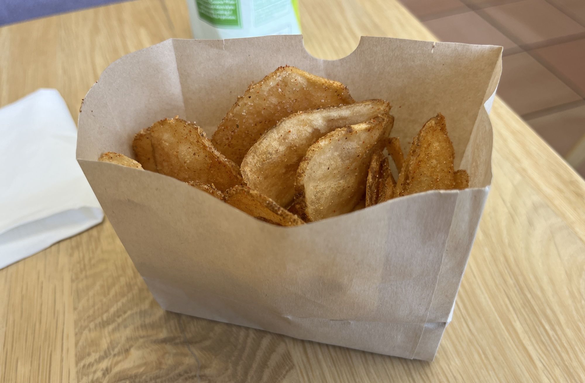 Brown paper takeout bag filled with seasoned potato wedges on a wooden table; a white napkin sits nearby and a green-labeled drink cup is blurred in the background.