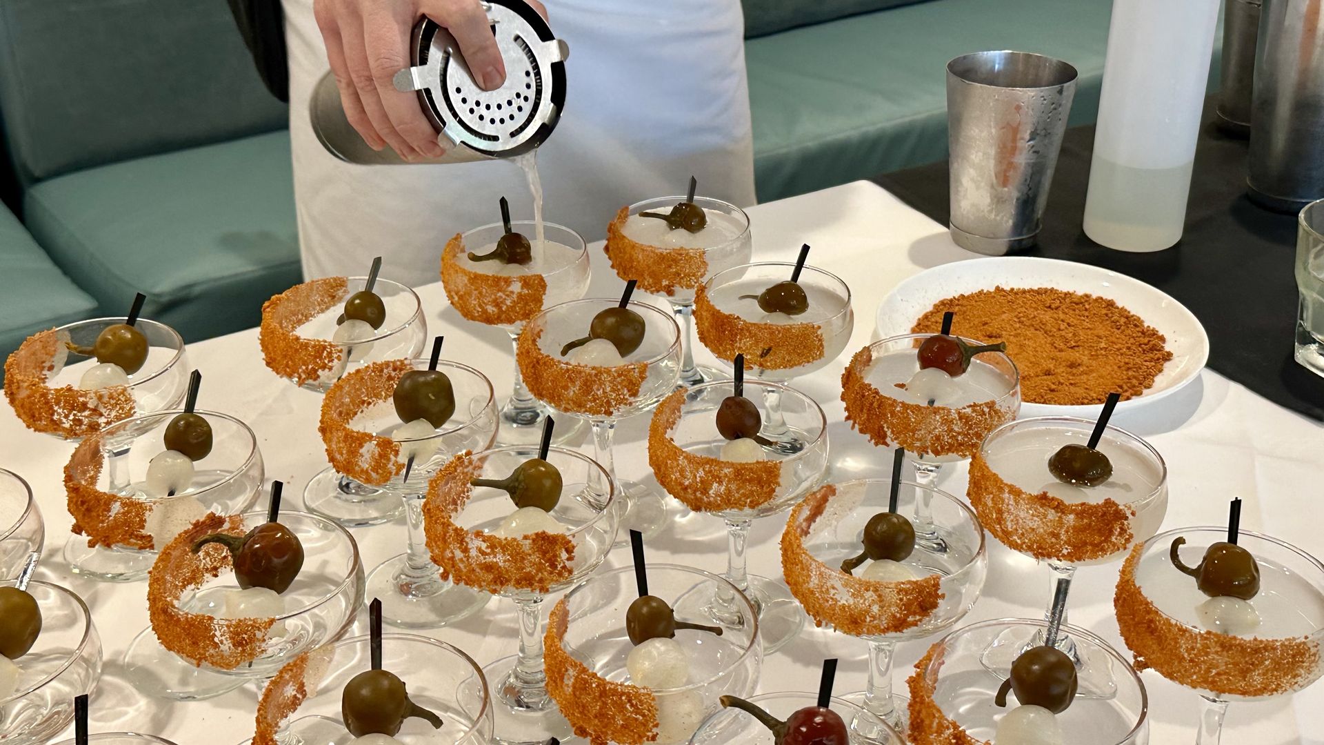 Bartender pouring clear liquid into cocktail glasses rimmed with orange seasoning, each garnished with green olives and translucent spheres on a white tablecloth.