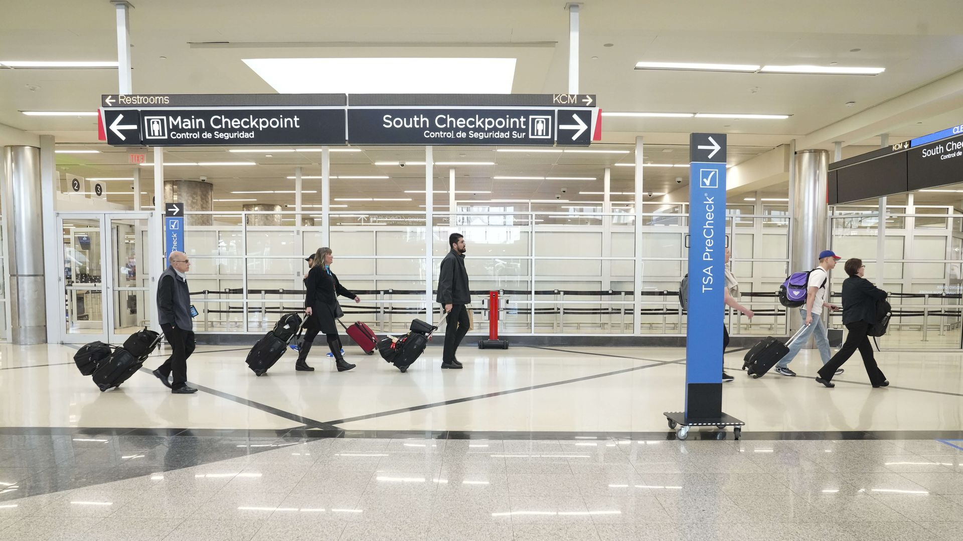Travelers carrying luggage walk through an airport. Signs with arrows pointing to the left for main check point and to the right for south check point are suspended from the ceiling. A blue and black TSA pre-check sign with an arrow pointing to the right is also visible.