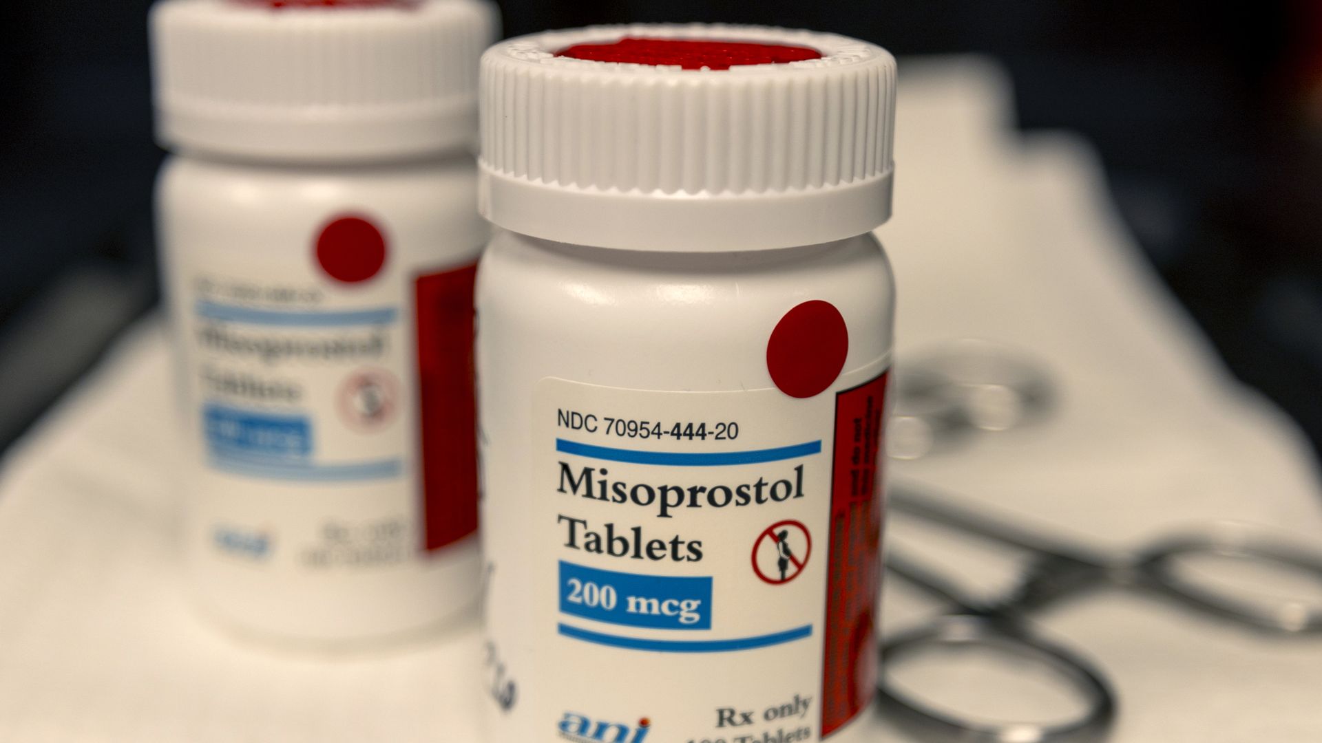 Close-up of white prescription bottles labeled Misoprostol Tablets 200 mcg and Mifepristone Tablets, with medical scissors in the background on a white surface.