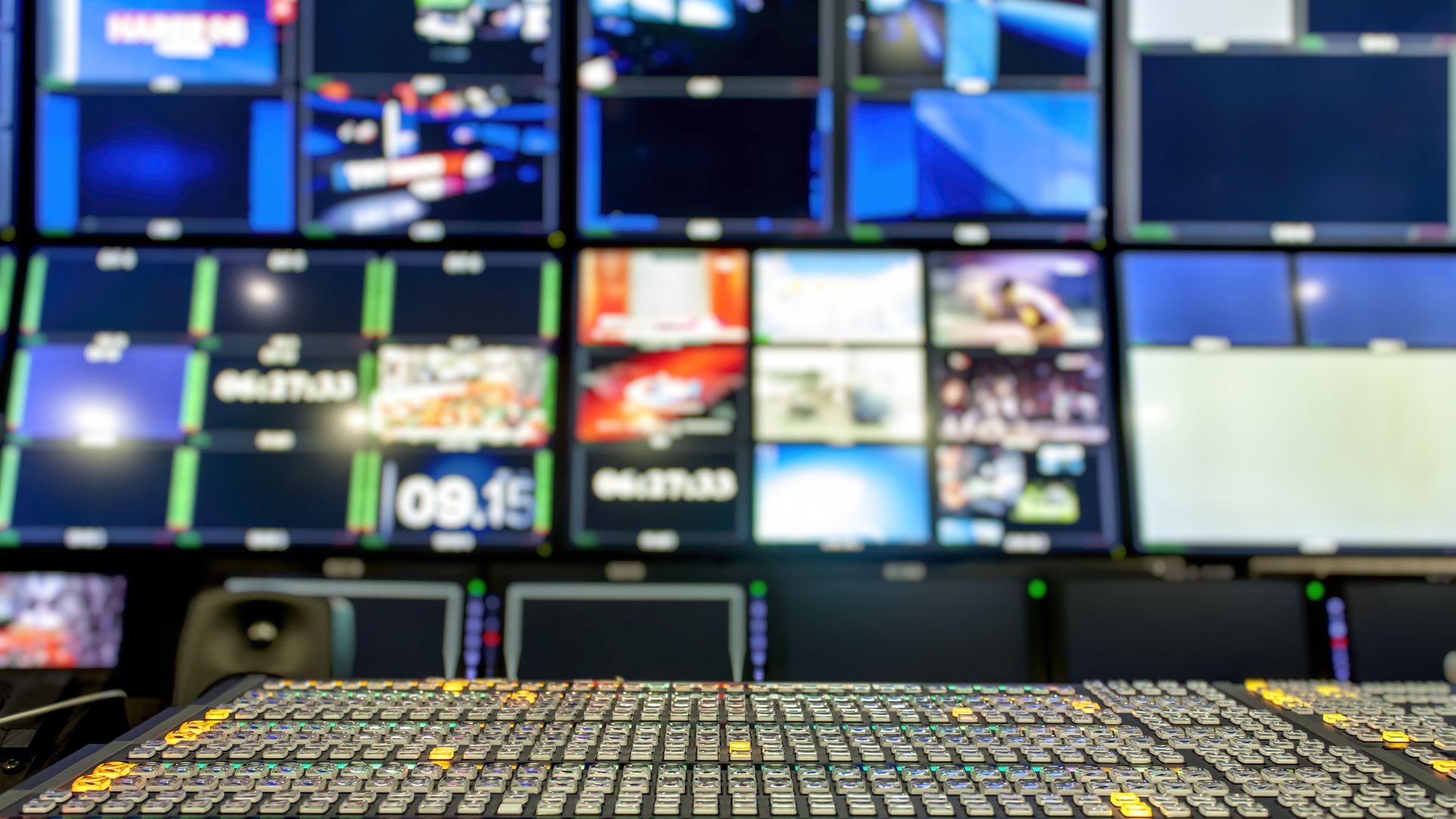 In this image, multiple rows of TV screens are in the background of a control panel. 