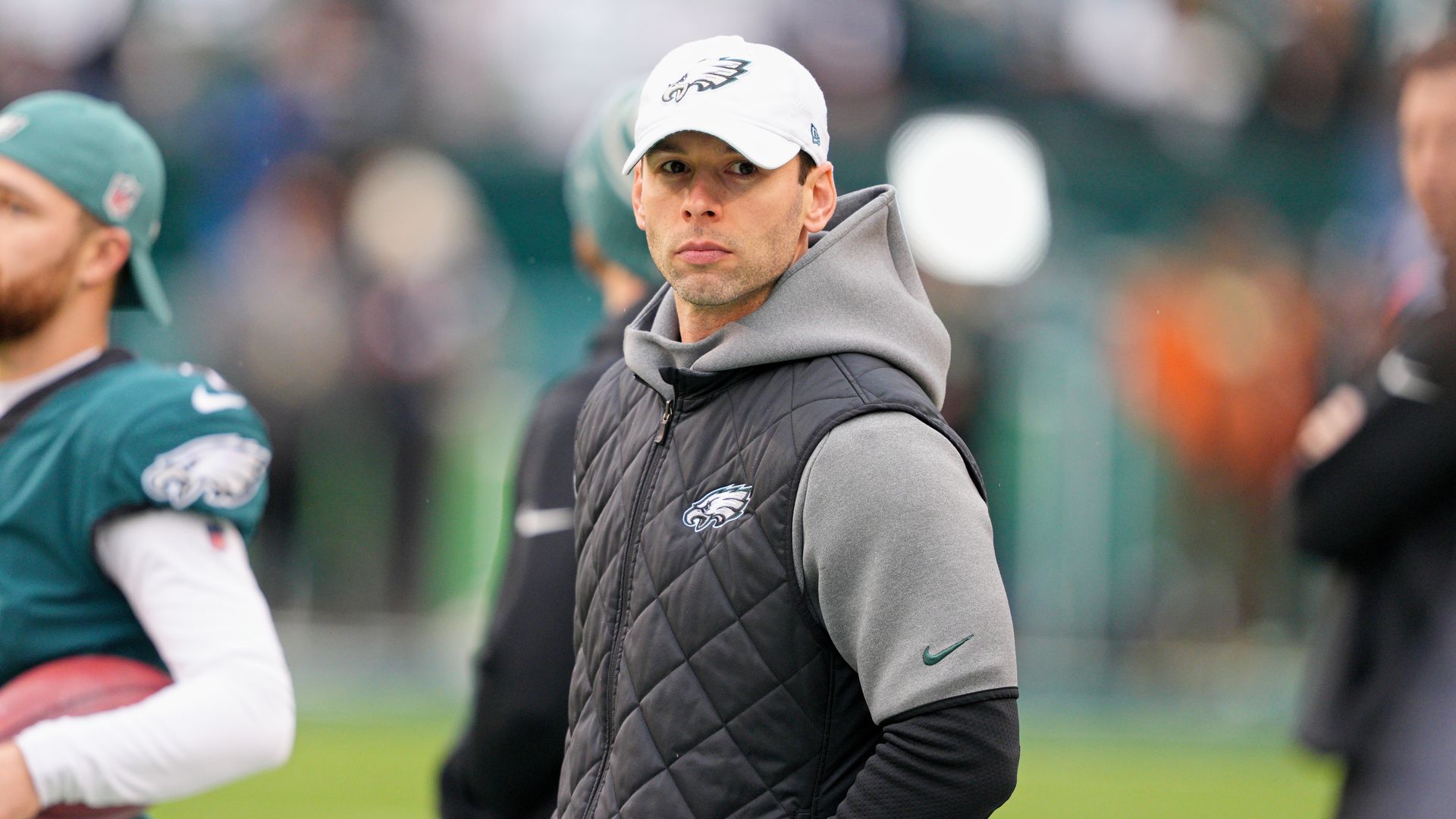 Jonathan Gannon stands on a football field wearing a white Philadelphia Eagles hat and a black and gray hooded jacket.