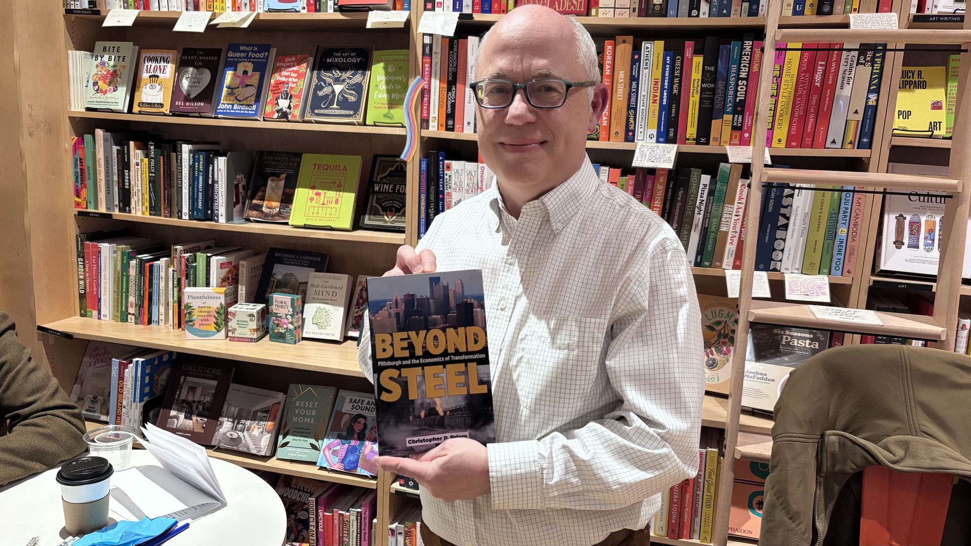 Smiling man with glasses in a white checkered shirt holding the book "Beyond Steel" in a colorful bookstore filled with various books on wooden shelves.