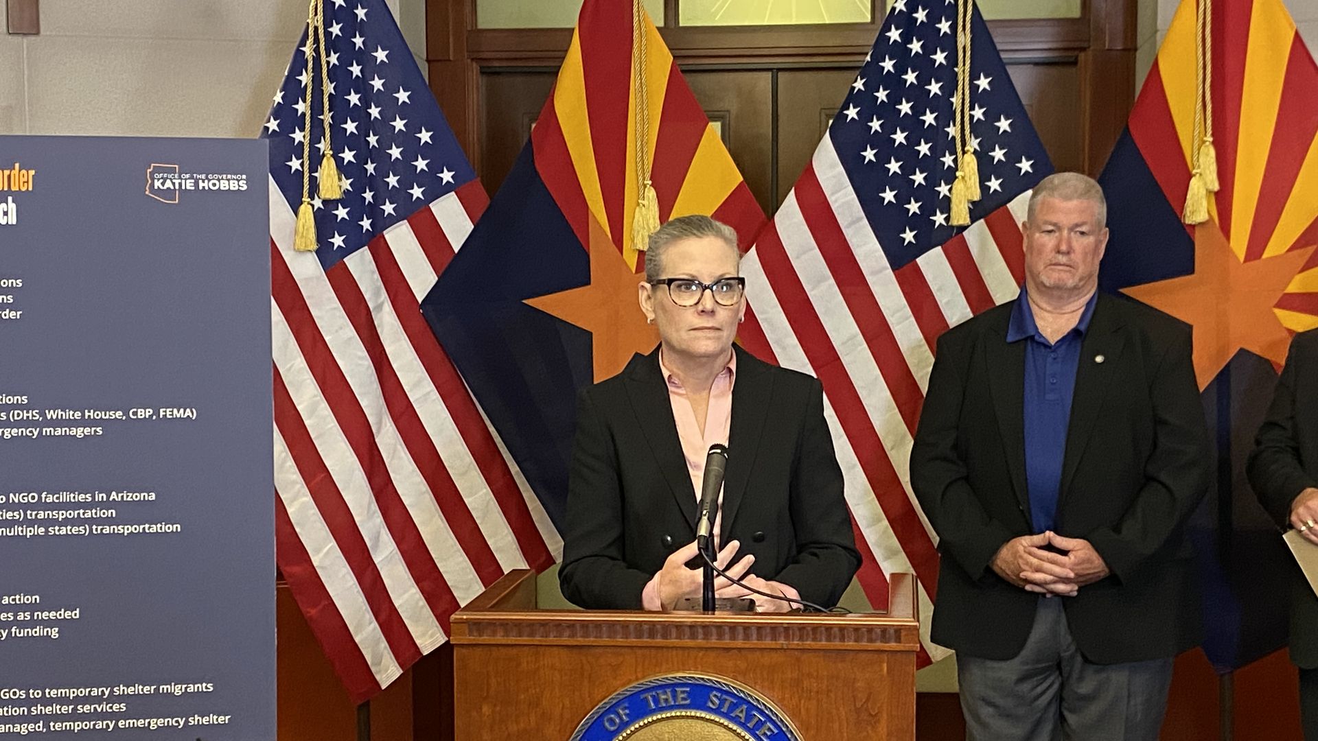 A woman stands at a lectern with a microphone in front of United States and Arizona flags. 