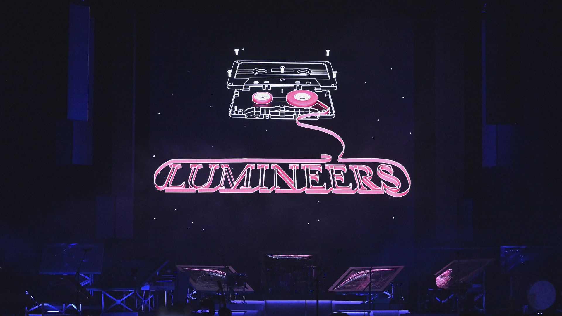A pink neon sign with a cassette tape trailing into the spelling out "Lumineers" at a concert venue.
