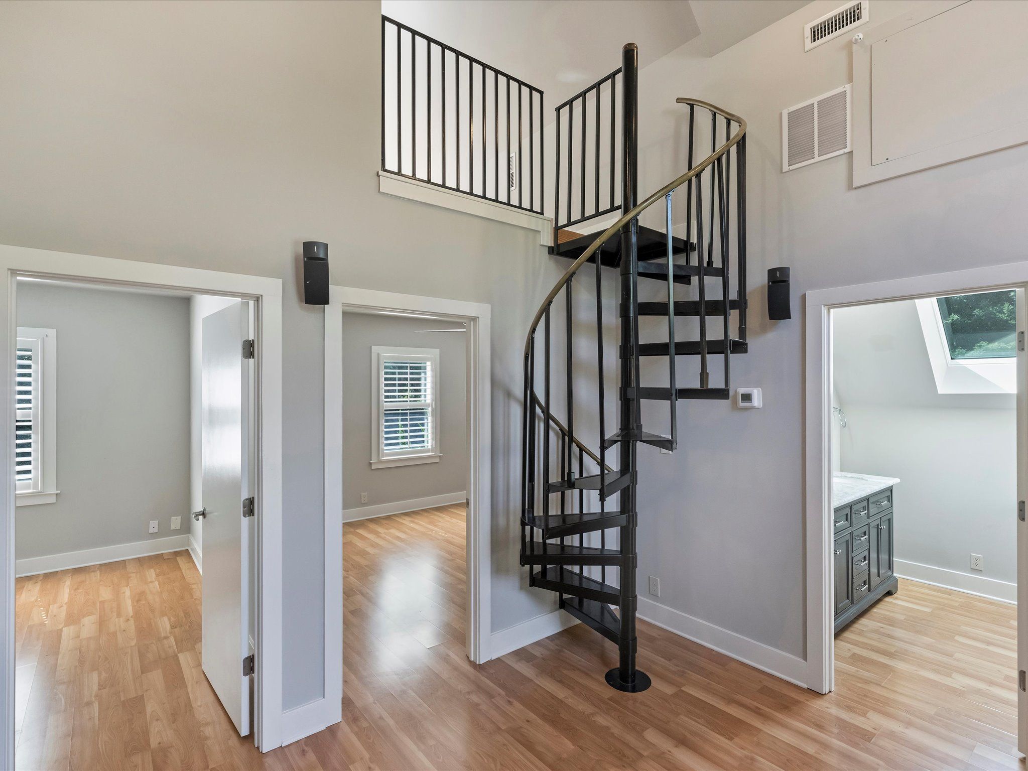 Interior view of a modern home with light wood floors, gray walls, a black metal spiral staircase, and three open doorways leading to rooms with windows and white trim.