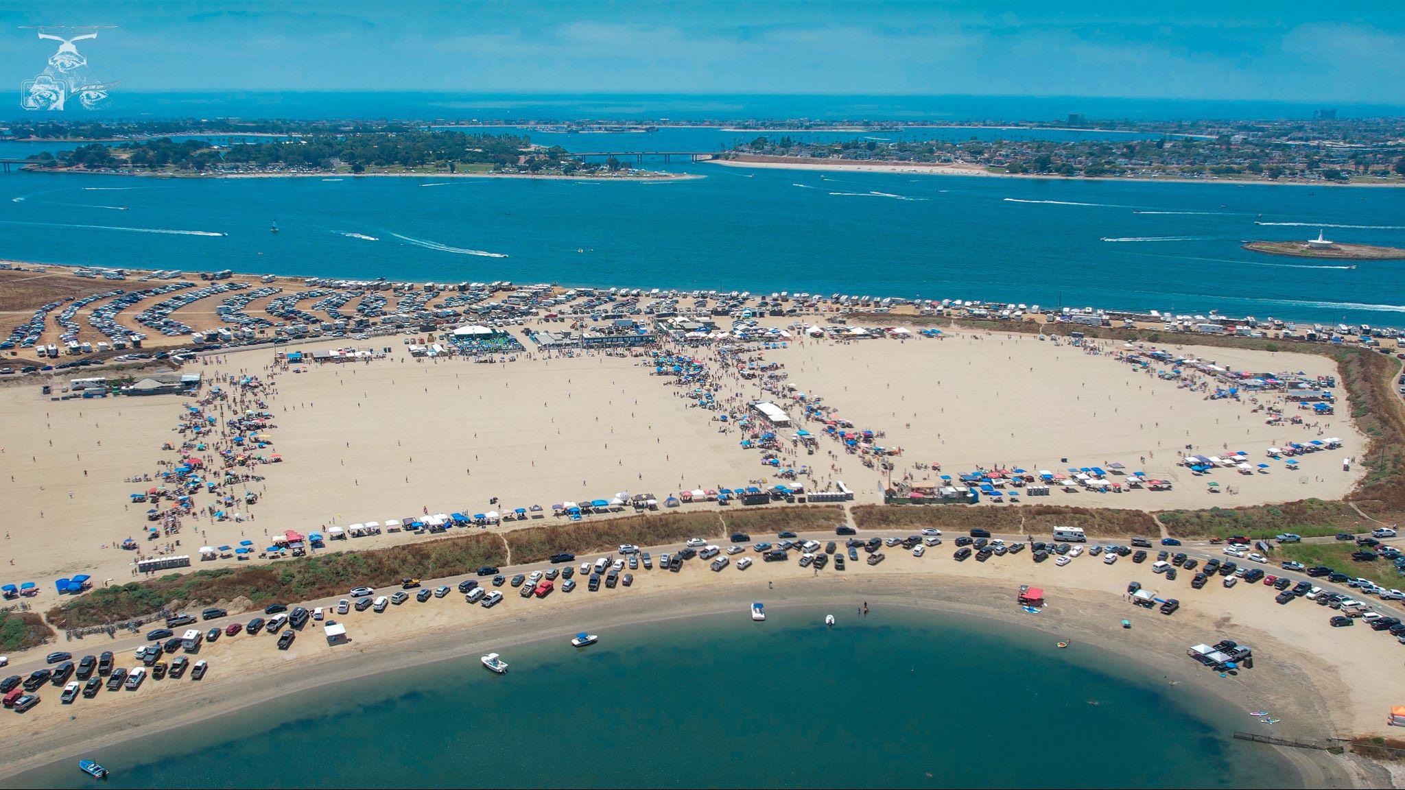 Thousands of people, cars and campers are lined up on a sandy beach of an island in the San Diego Bay forming playing fields for a large tournament of a beach game called Over The Line.