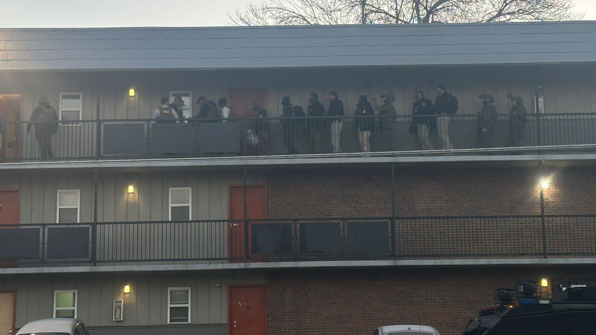 Several men in jackets and military gear stand inside an outside hallway at an apartment complex. 