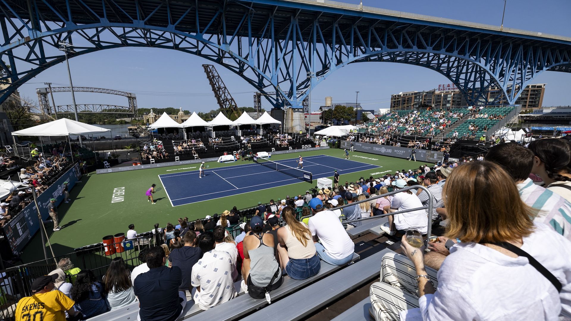 Crowd watching a tennis match on a blue and green court under a large blue bridge on a sunny day, with tents and seating in the background and players in action on court.