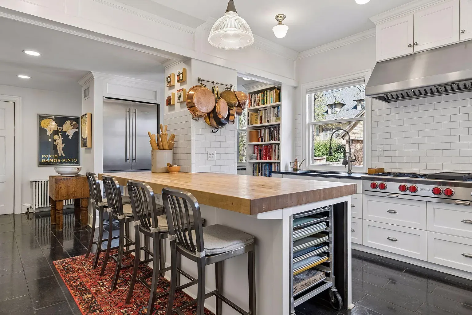 Bright white kitchen with a large wooden island, four metal stools, copper pots hanging, stainless appliances, white subway tile, and a bookshelf by the window.