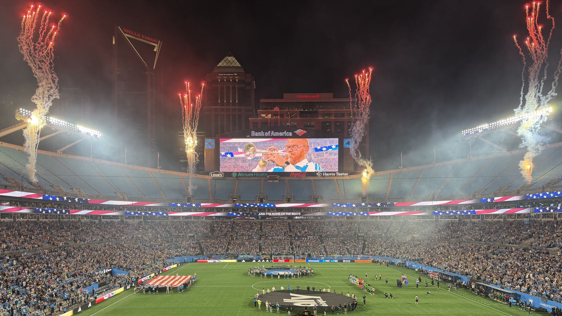 Night stadium filled with fans watching a patriotic pre-game ceremony; fireworks erupt, an American flag is displayed on the field, and a video screen shows a man playing the trumpet.