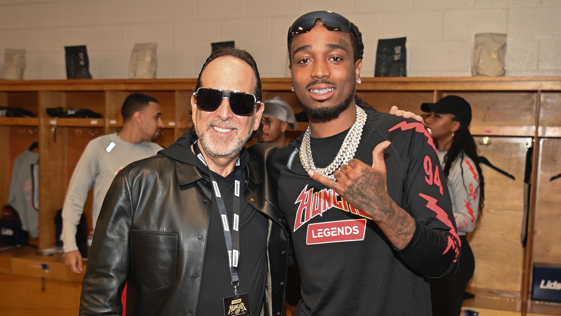 A man wearing dark sunglasses and all black clothing poses with another man in a locker room