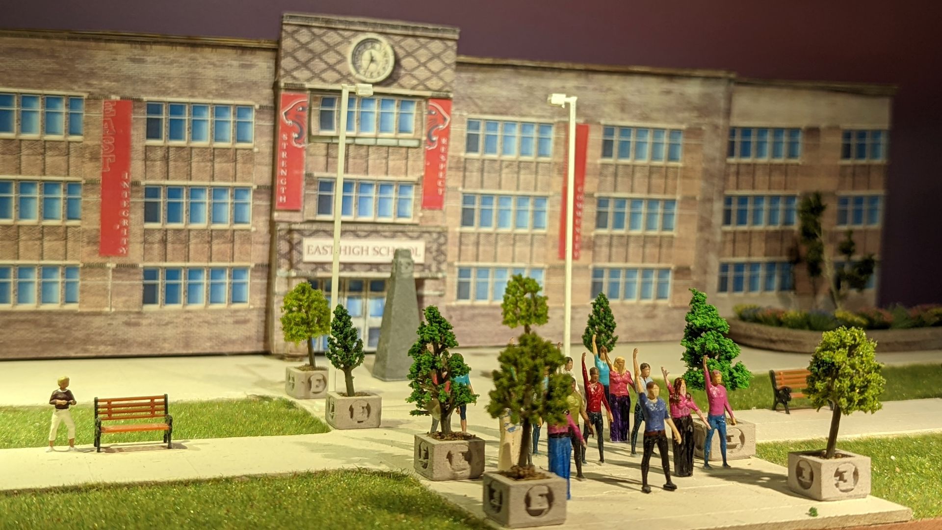 Human miniature figures pose outside of a miniature high school in a diorama.