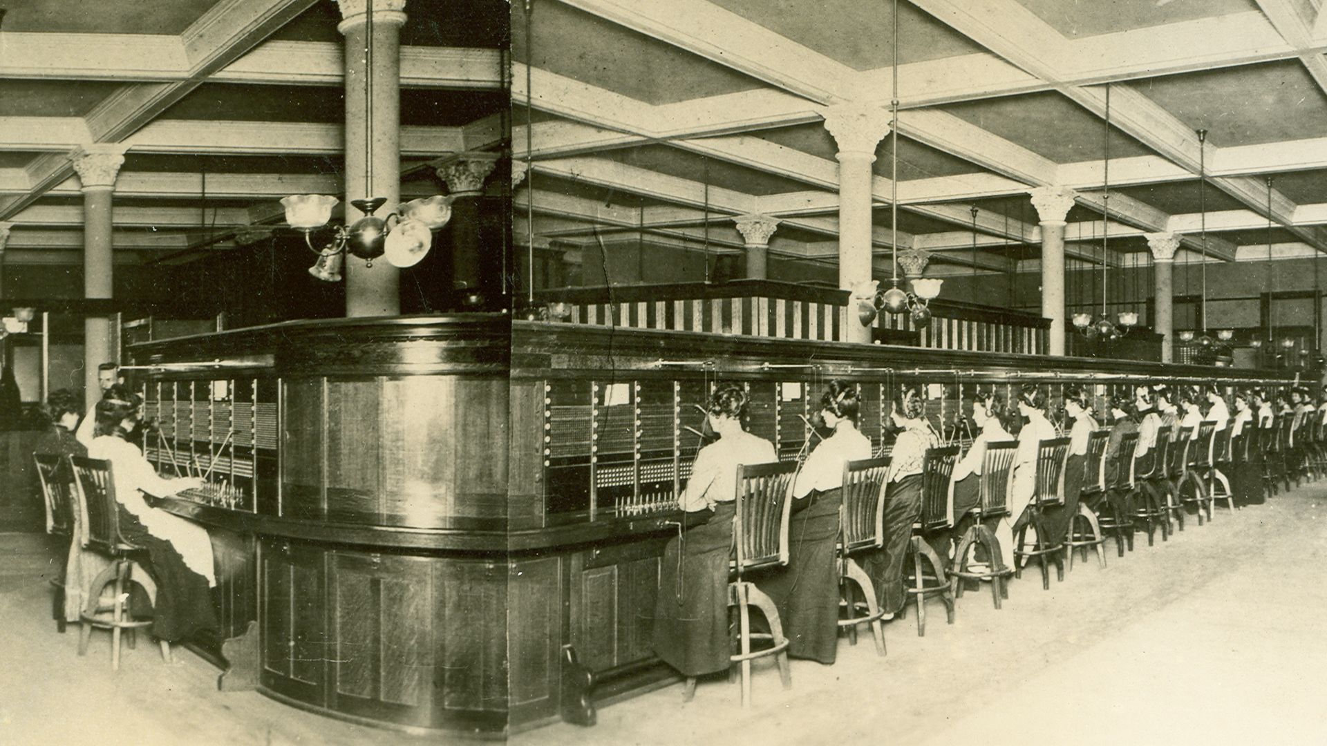 Black-and-white photo of a vintage switchboard room with a long row of operators at desks, a large central wooden counter, tall columns, and hanging lamps.