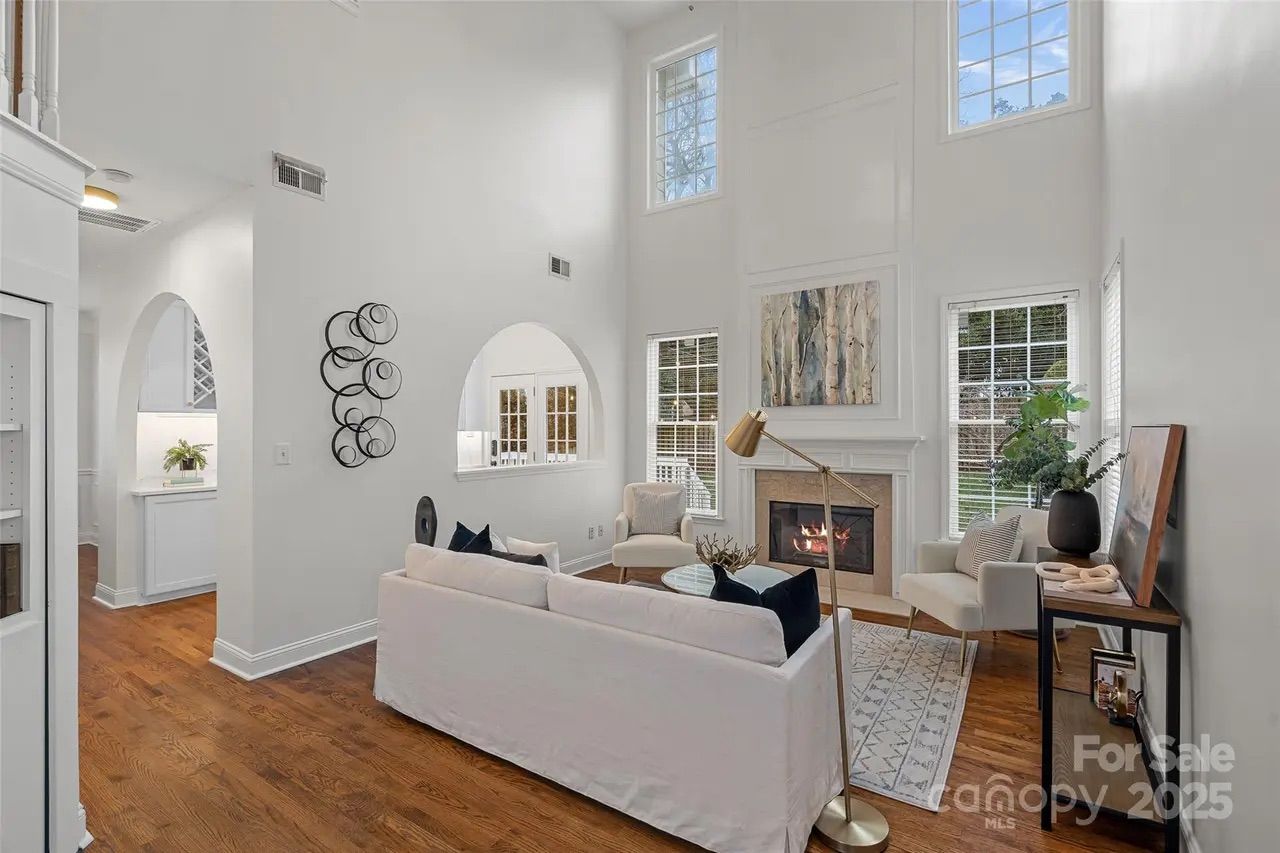 Bright living room with white walls, wooden floor, white couch, two armchairs, a glass coffee table, a fireplace with fire, and tall windows letting in natural light.