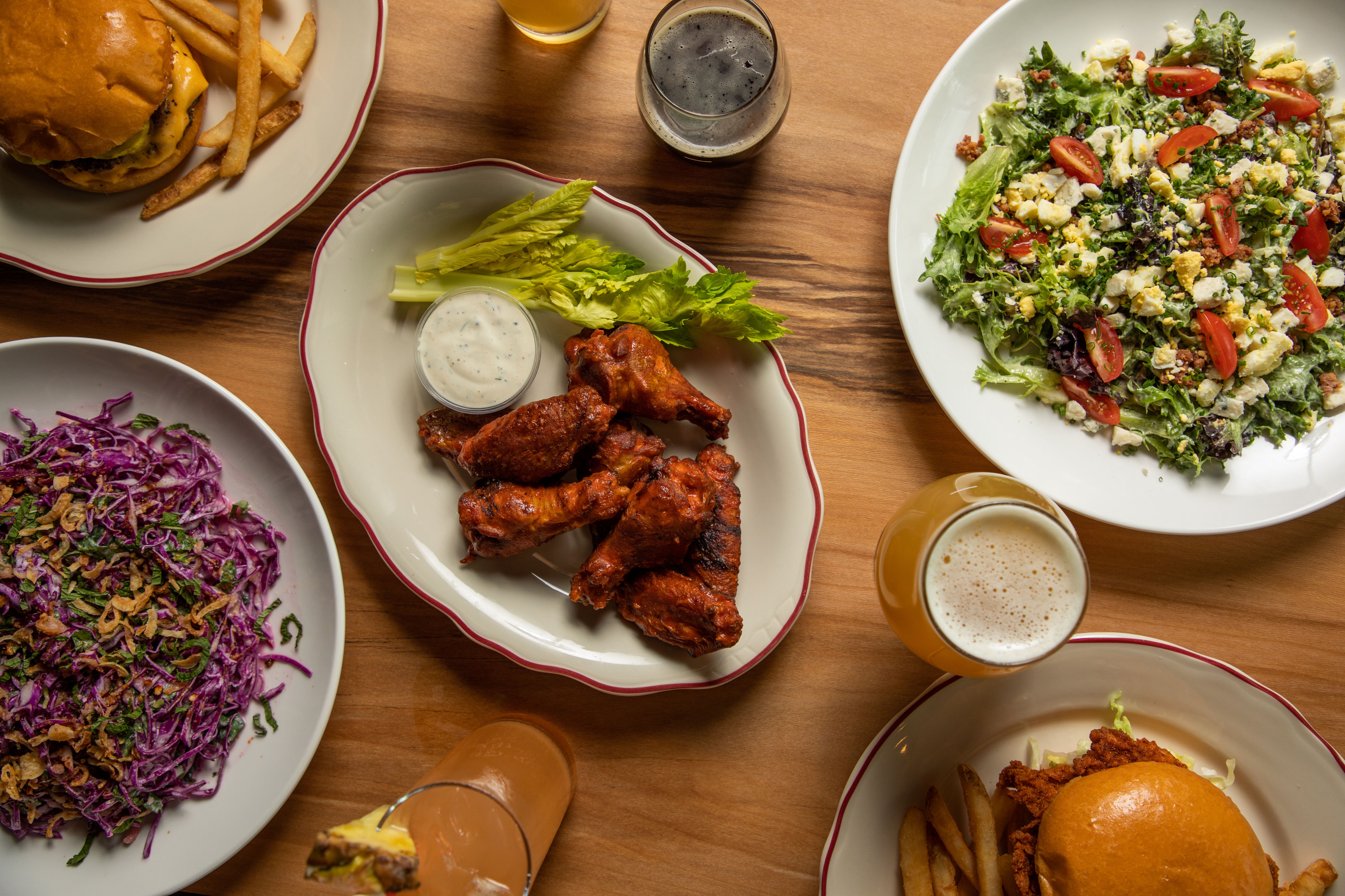 A full wooden tabletop covered in salads, chicken wings and beers is viewed from above. 