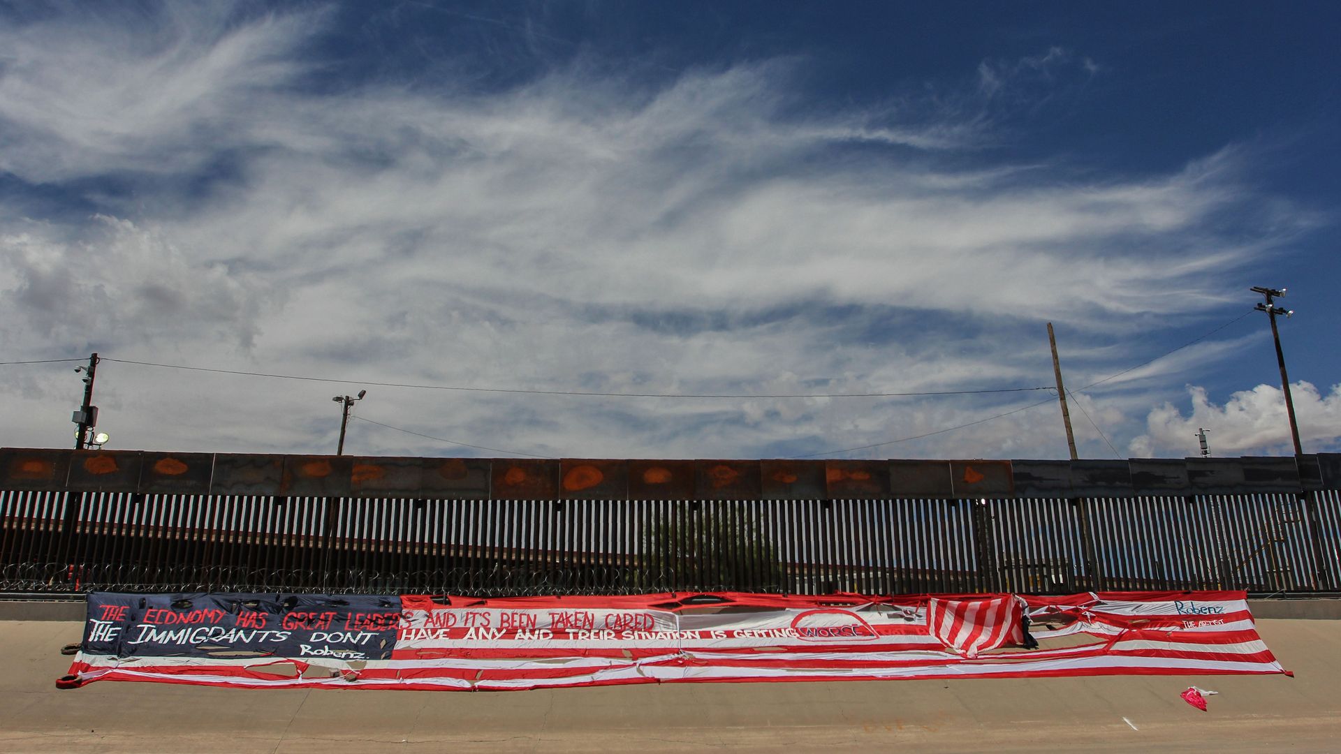 This image shows a larger horizontal American flag in front of a wall at the southern border.