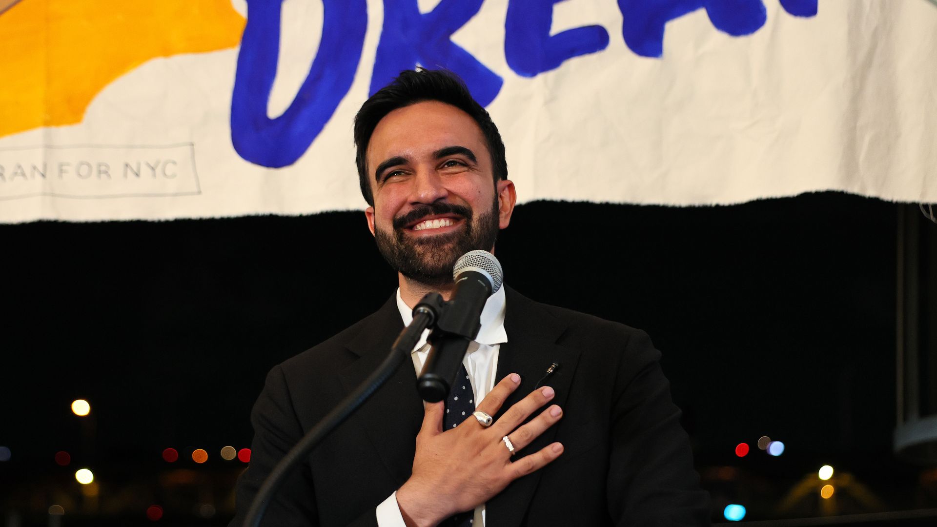 Zohran Mamdani, wearing a black suit with a hand on his chest, standing in front of a white banner and a black sky.