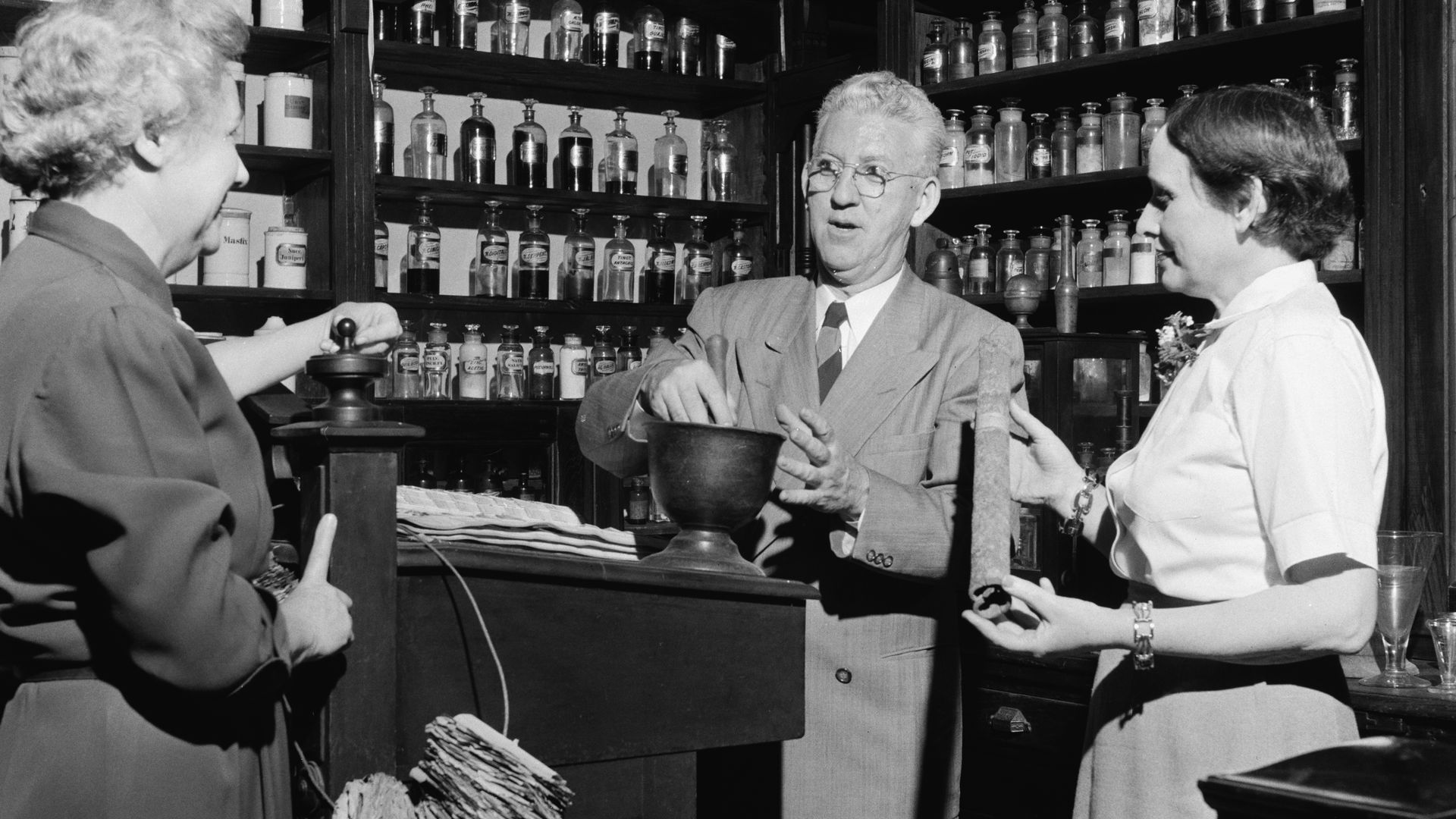 A black and white historical photo of three people gathered in an apothecary-like space as a man mixes things in a large bowl.