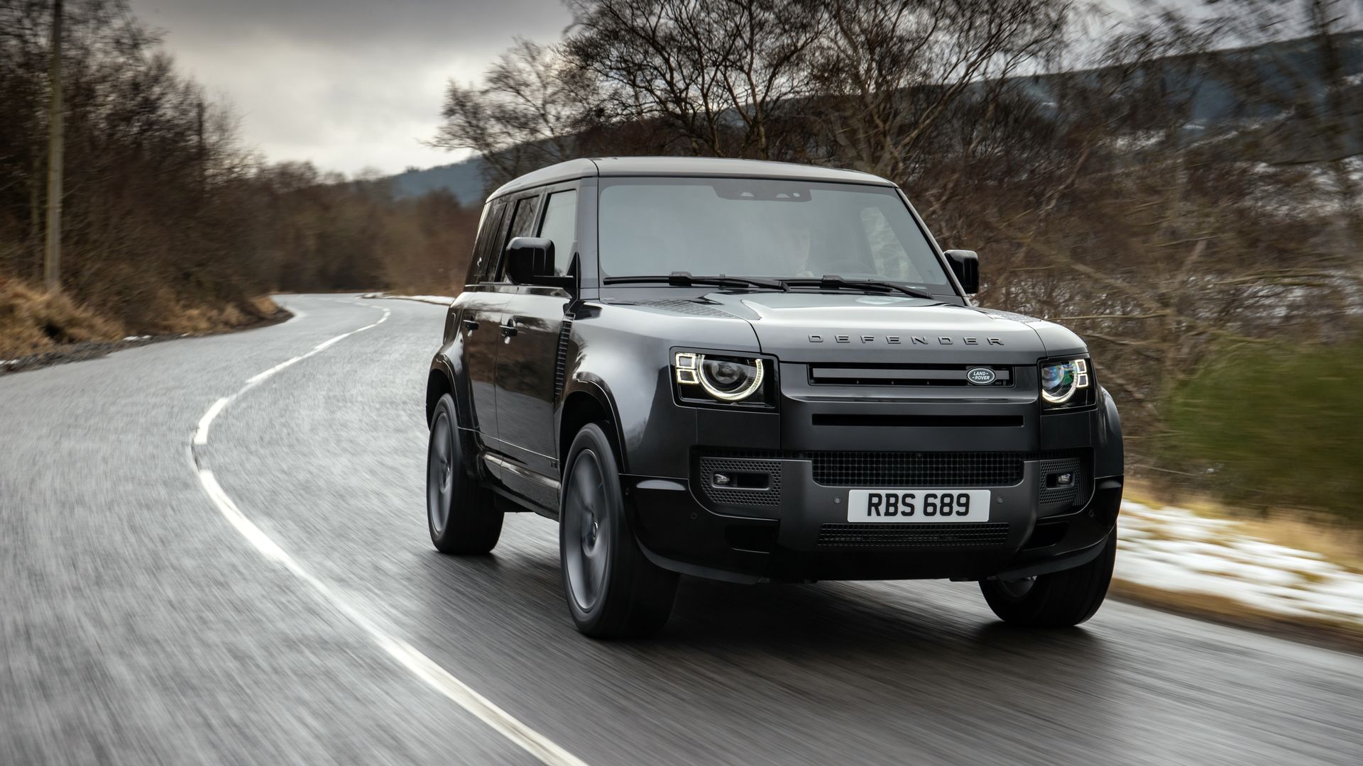 A silver Land Rover Defender drives along a winding, wet road under a gray, overcast sky; leafless trees line the roadside, with motion blur conveying speed.