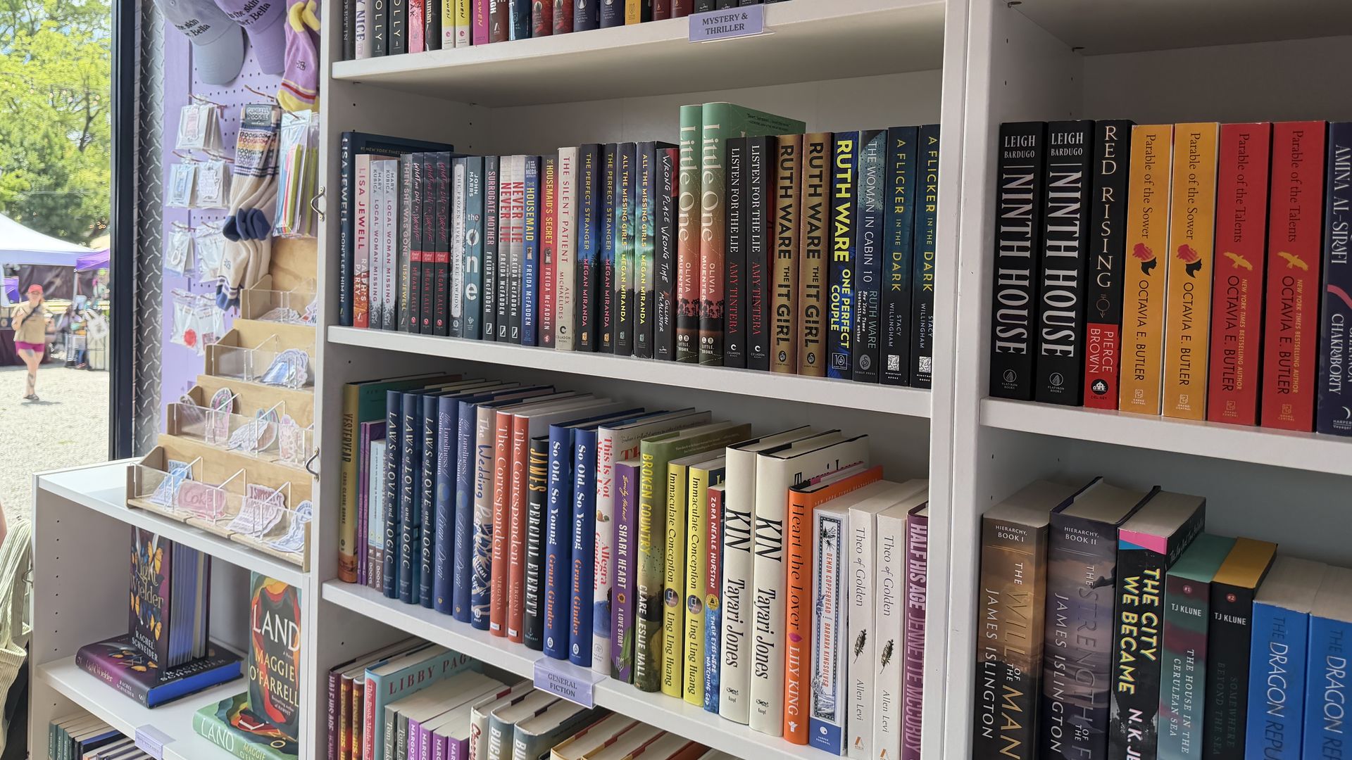 Interior of a bookstore with white shelves packed with colorful book spines, labeled 'Mystery & Thriller.' A window on the left reveals trees outside and hanging hats.