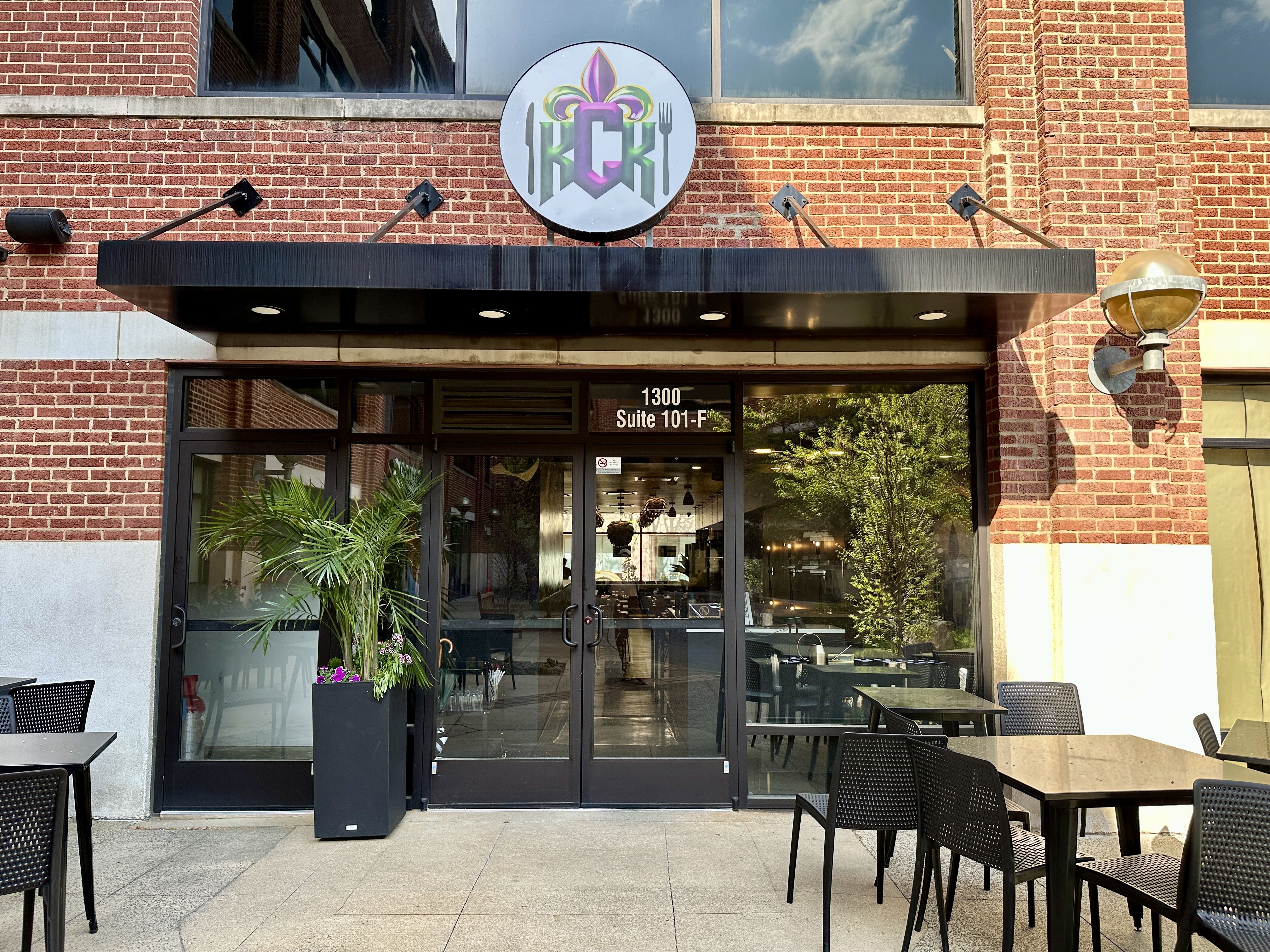 Brick storefront with a black awning and glass doors. A round, colorful sign above. Outdoor seating with black tables and chairs, a potted palm, and tree reflections in the glass; address 1300 Suite 101-F.