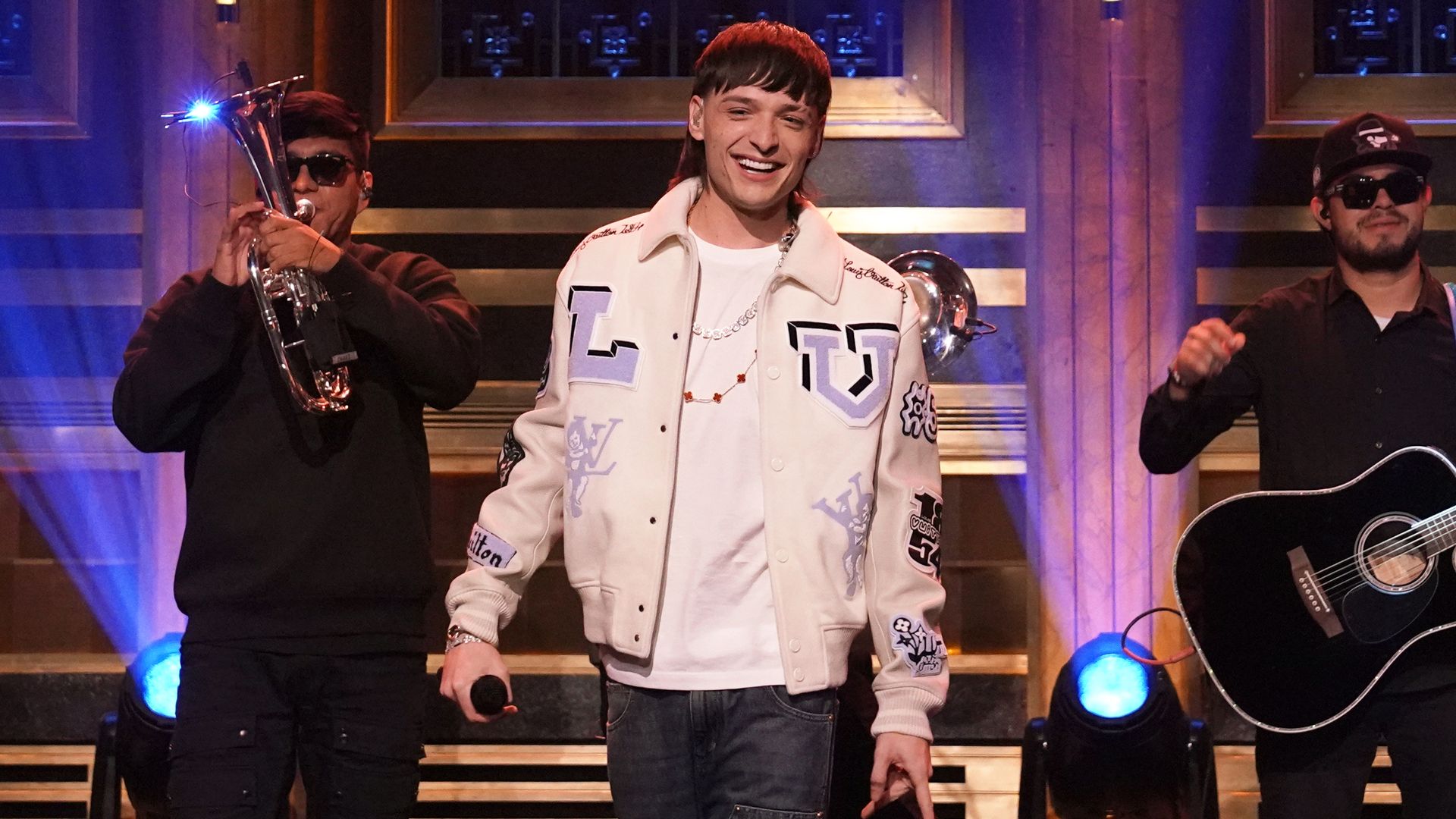 Peso Pluma smiling on stage with a band behind him. 