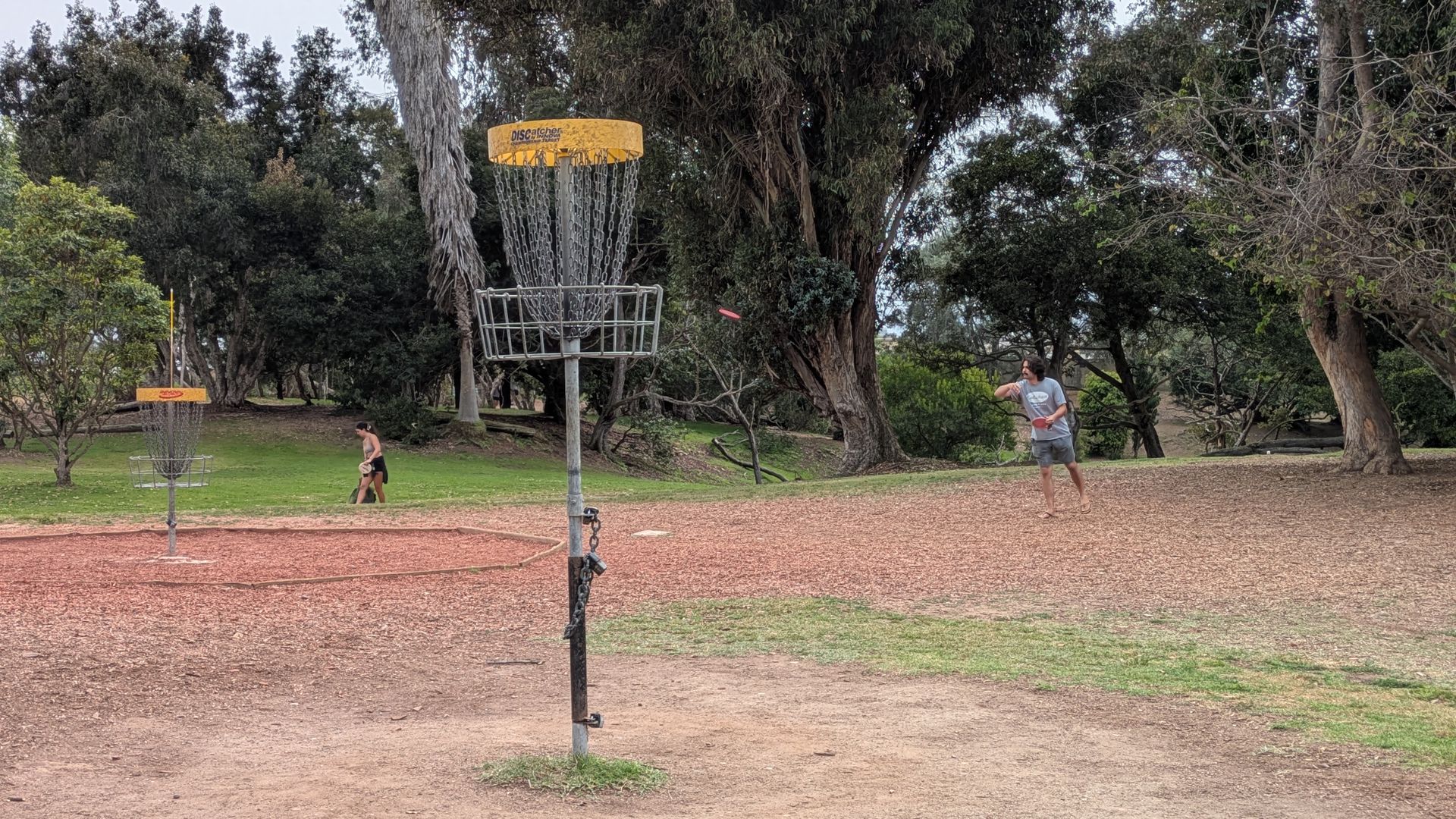 Disc golf park with two players; a yellow-topped basket in the foreground and a pink disc mid-flight toward a man in gray shirt and shorts on the right, while another person walks left.
