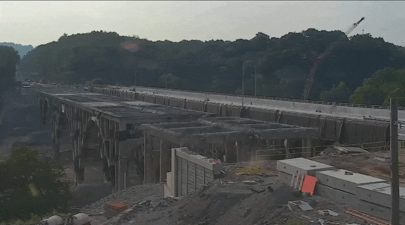 A highway bridge exploding and releasing a cloud of dust on an empty highway with green hills in the background.