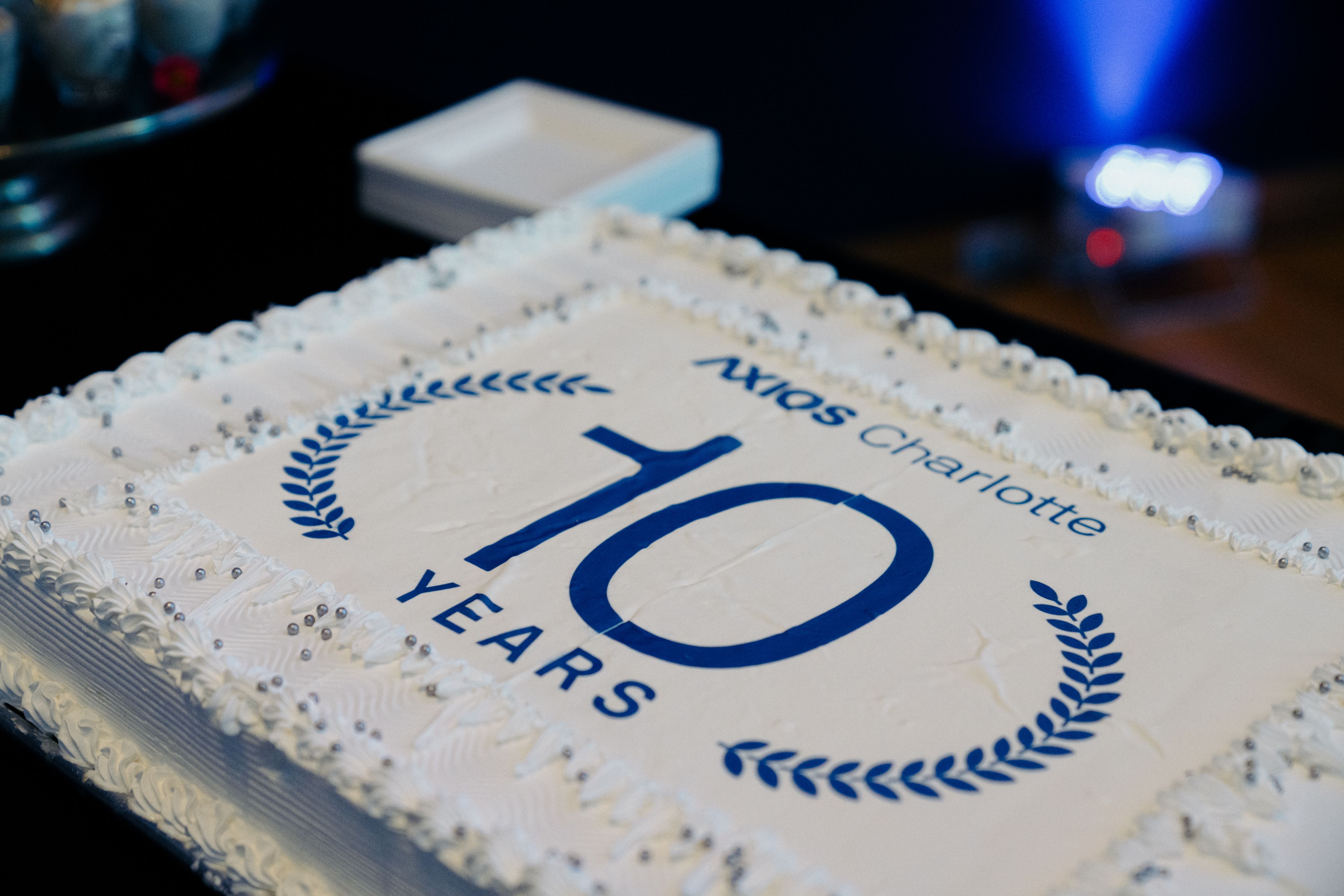 Rectangular white cake with decorative edges and silver beads, printed with blue text "AXIOS Charlotte 10 YEARS" surrounded by laurel branches, celebrating a 10-year anniversary.