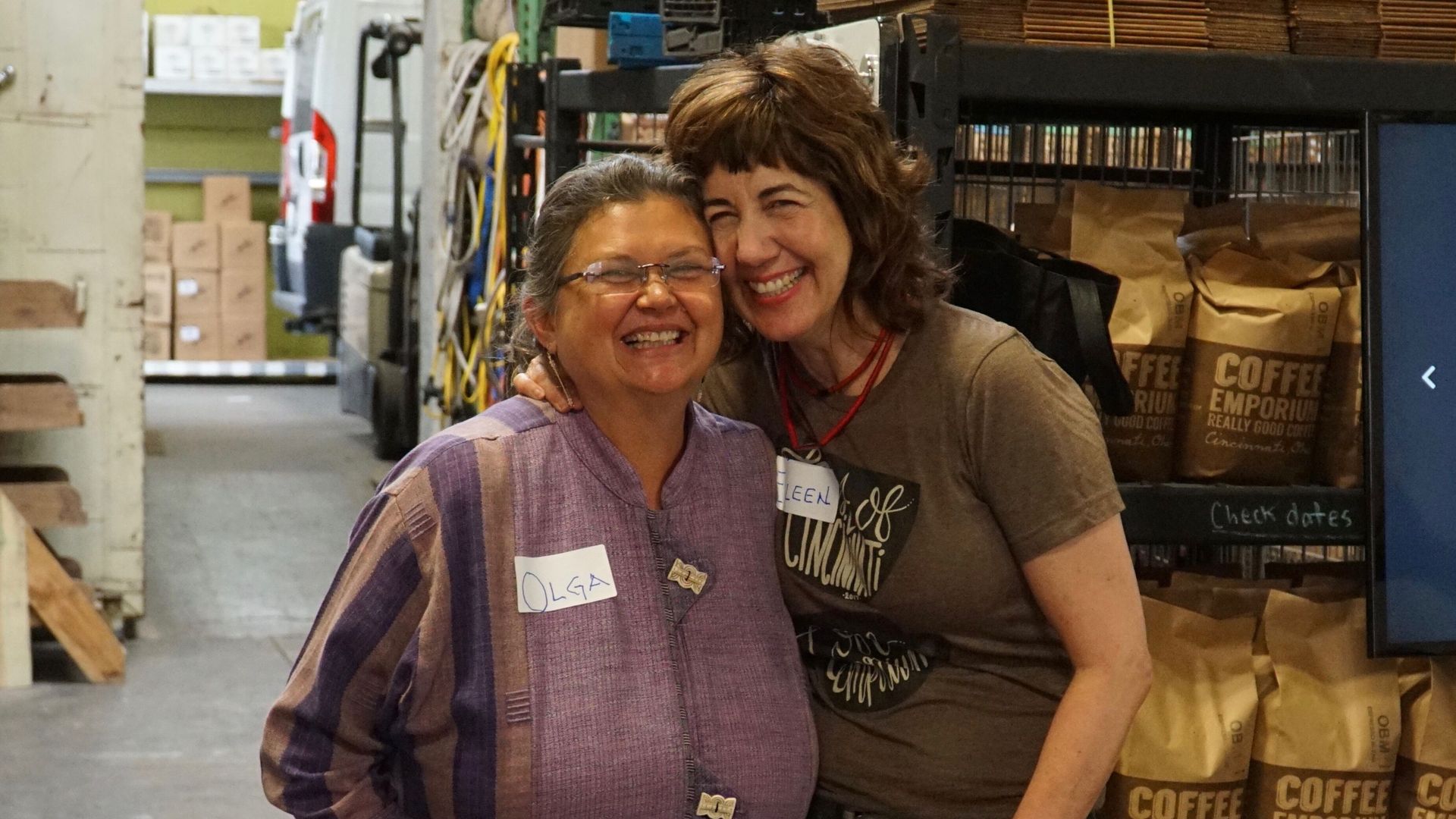 Two women smile and hug in a cluttered warehouse aisle, wearing name tags. One in a purple plaid shirt, the other in a brown tee, with shelves, boxes, and coffee bags in the background.