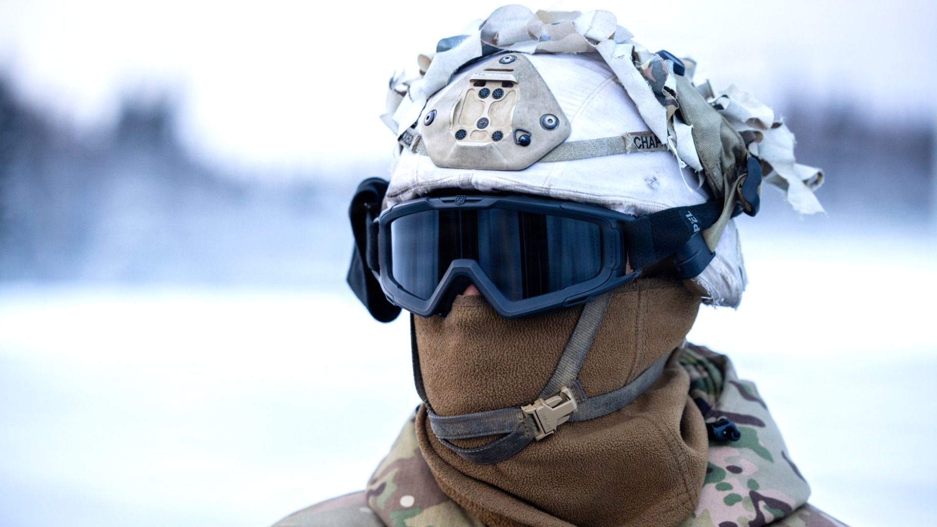 Close-up of a soldier wearing winter camouflage uniform, white helmet with fabric strips, black goggles, and brown face covering against a snowy background.