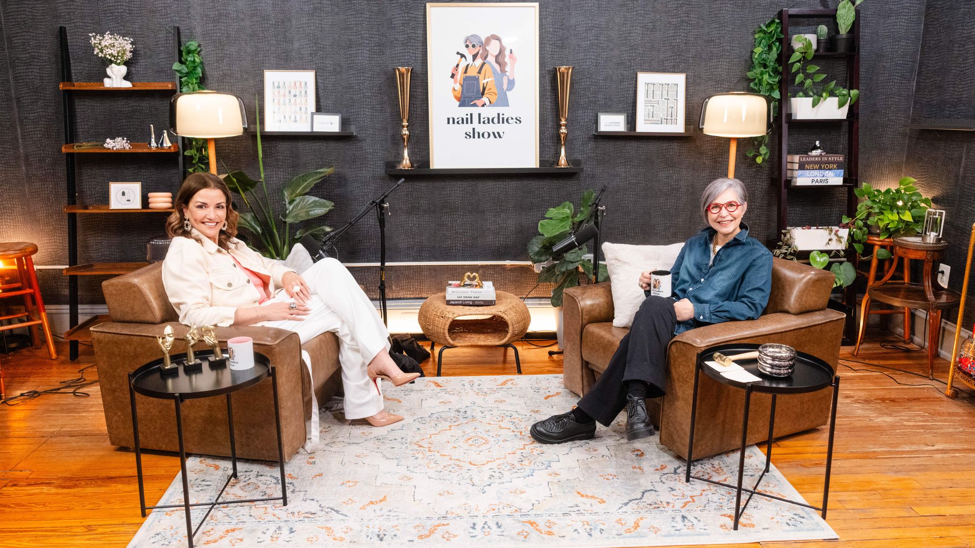 Two women sitting in brown armchairs in a studio with dark walls, plants, a patterned rug, and a framed "nail ladies show" poster behind them. They smile at the camera.