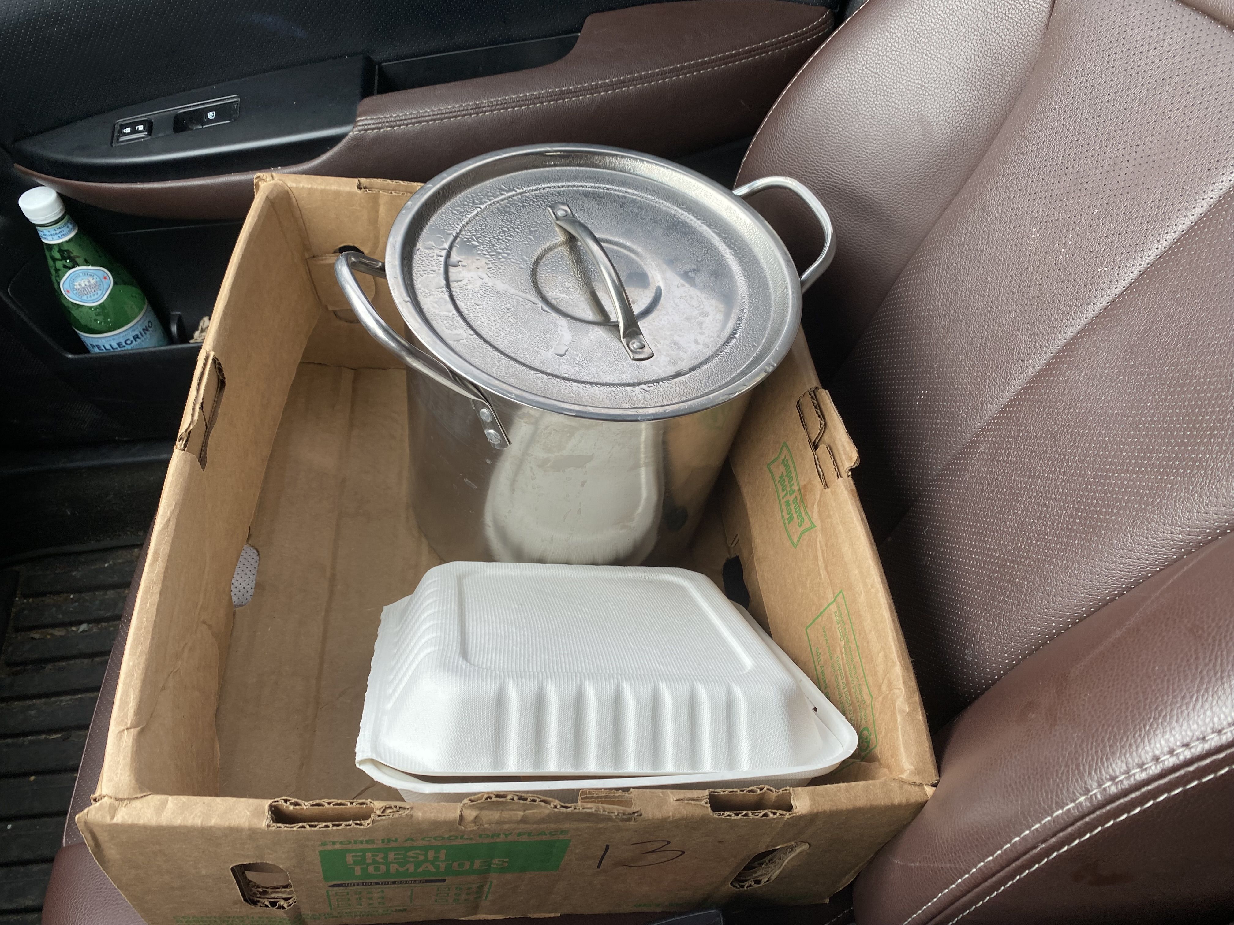 A stainless steel pot next to a white takeout container, both inside a brown box on the seat of a car.