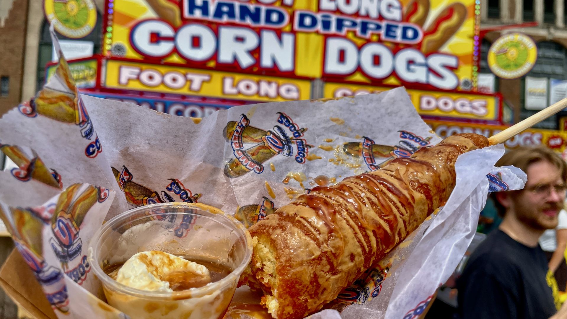 Corn dog on a stick with sauce, wrapped in paper, held up in front of a bright sign reading "CORN DOGS" and "FOOT LONG," with a blurred crowd in the background.