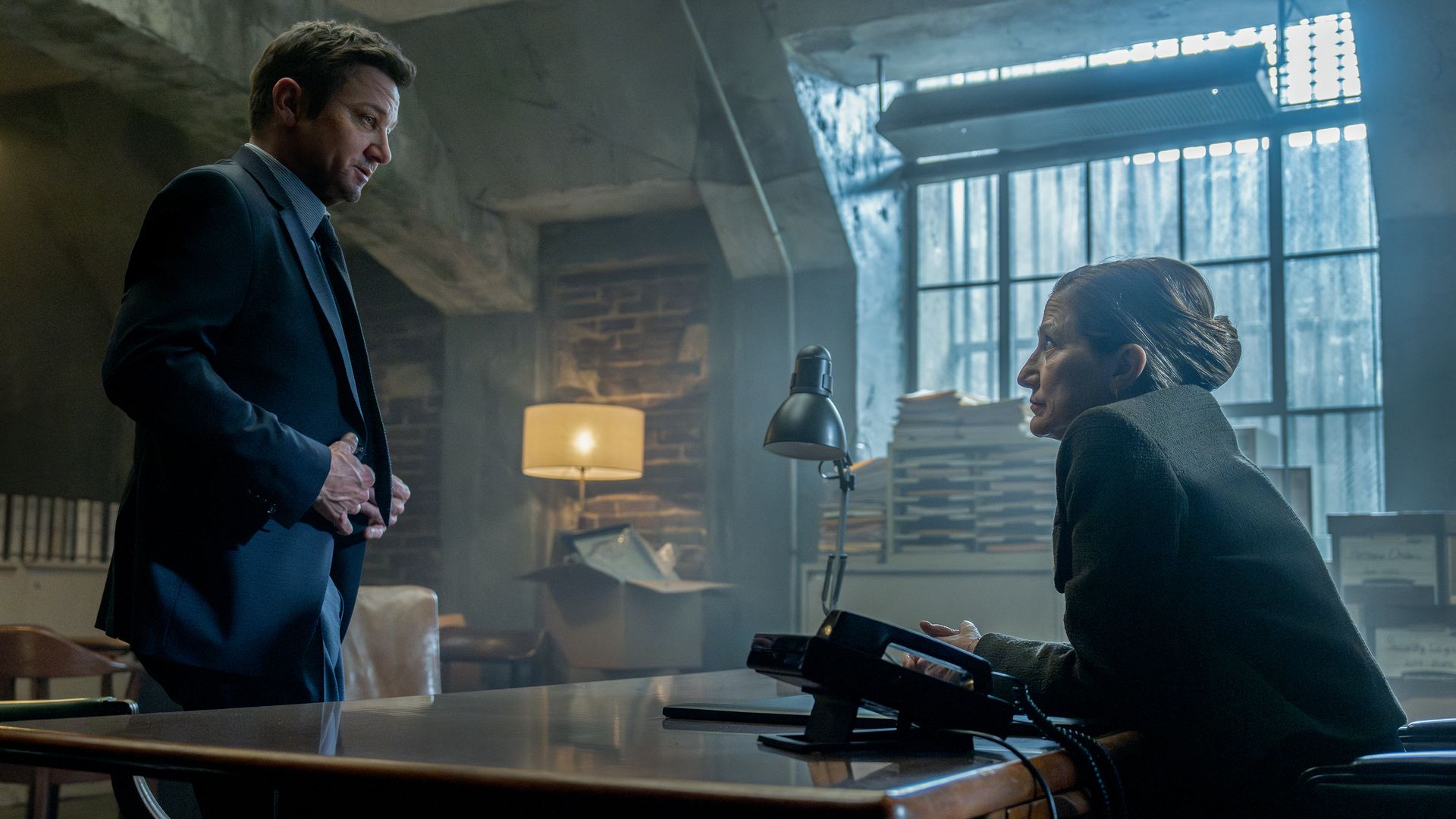 Man in a suit standing and woman sitting at a desk with a telephone in a dim, industrial-style room with concrete walls and a large window with metal bars.