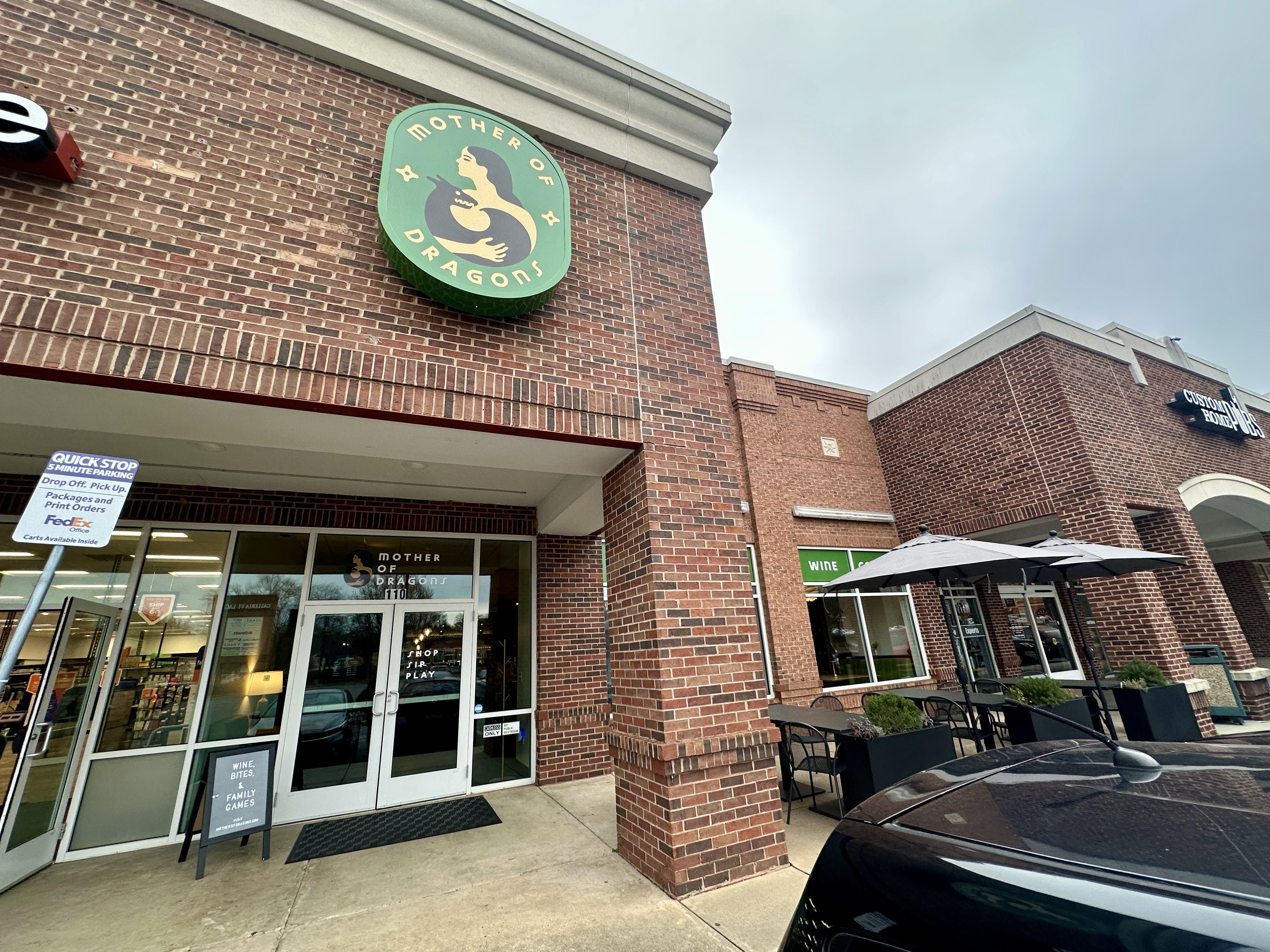 Outdoor view of a brick storefront with signs for "Mother of Dragons" featuring a woman holding a dragon, and a cafe area with tables and umbrellas under a cloudy sky.