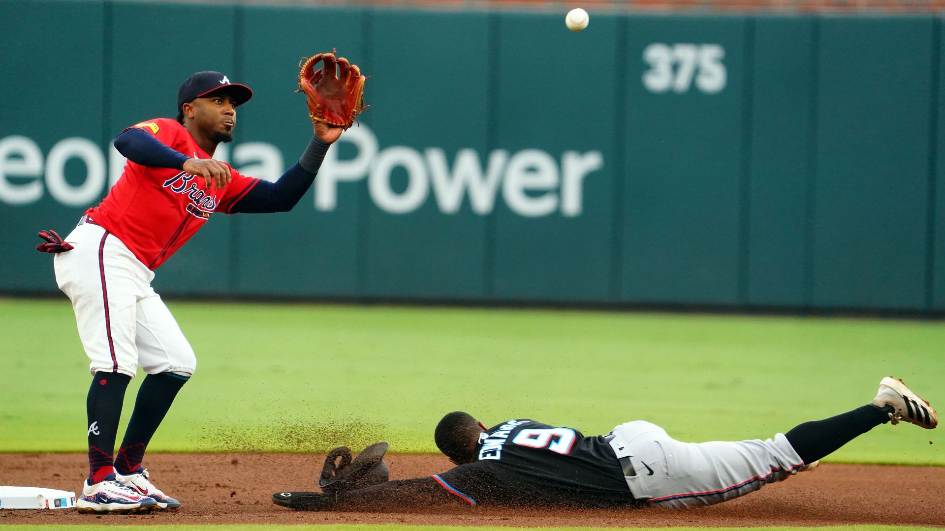 A baseball player wearing a black jersey and gray pants slides with hands outstretched toward a base. A player wearing a red jersey and white pants with red piping and his feet on the base is preparing to catch a ball.