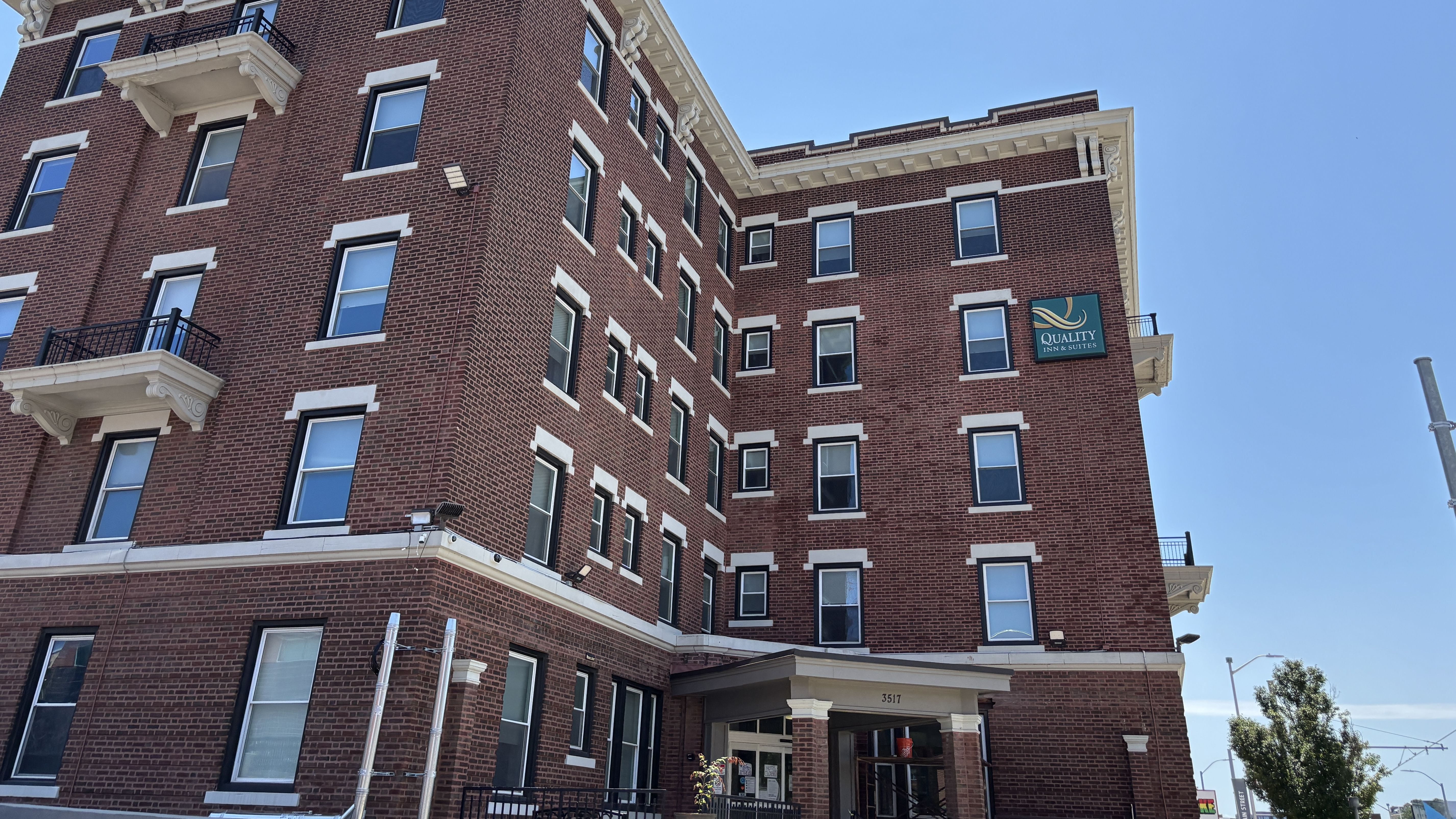 Red brick hotel building with white trim and decorative cornices, many windows and small balconies. A Quality Inn & Suites sign sits on the right, set against a clear blue sky.