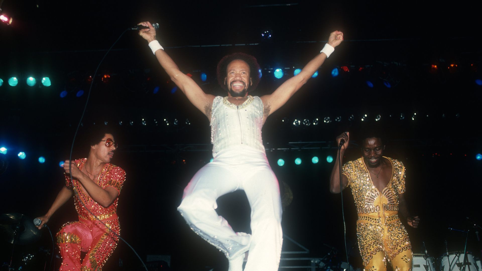 Center stage singer in a white sparkling outfit raises arms, flanked by red- and gold-sequined dancers; colorful stage lights illuminate a lively performance.