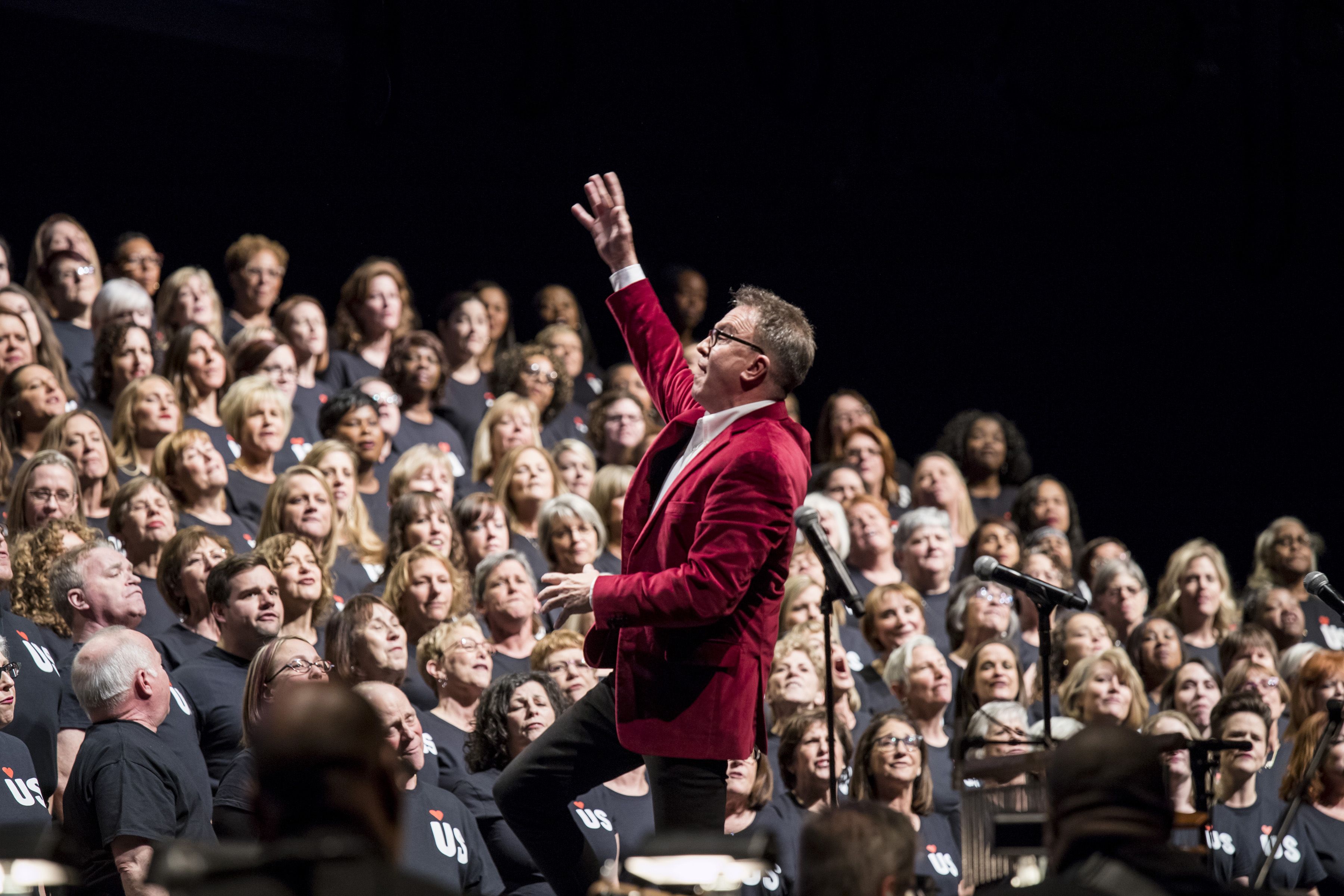 David Brown, in a red jacket, leads a large choir wearing black shirts, performing on stage with microphones and dark background.