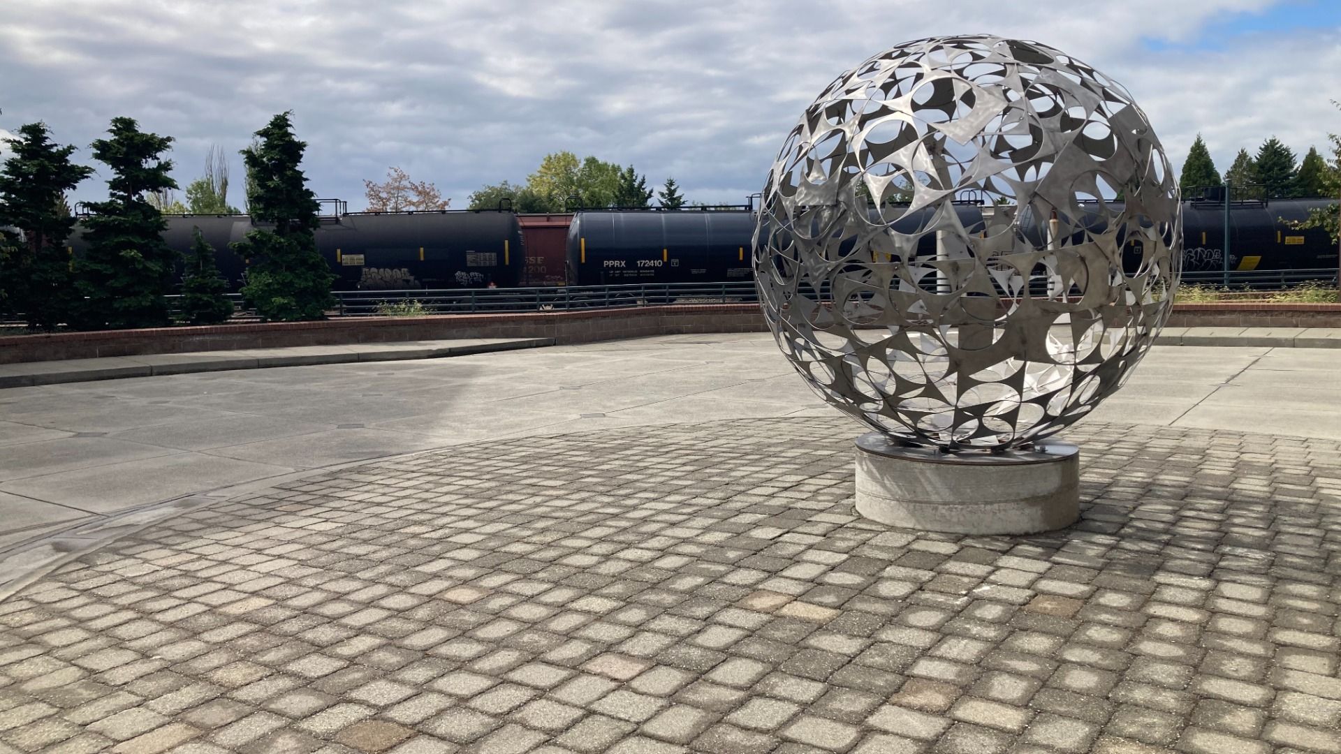 A hollow metal sculpture of a sphere on a stone  plaza in front of a train. 