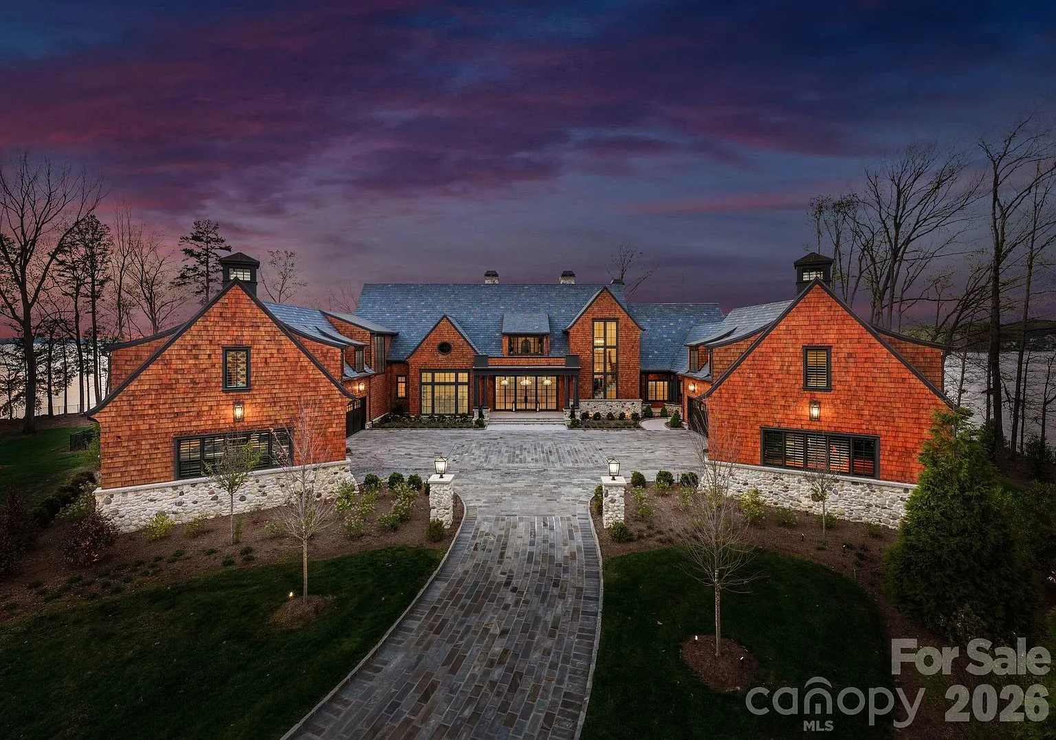 Luxurious red-brick estate at dusk with multiple gabled roofs, stone foundation, warm windows, and a curved stone driveway lining a manicured lawn, bare trees, and a twilight purple sky.