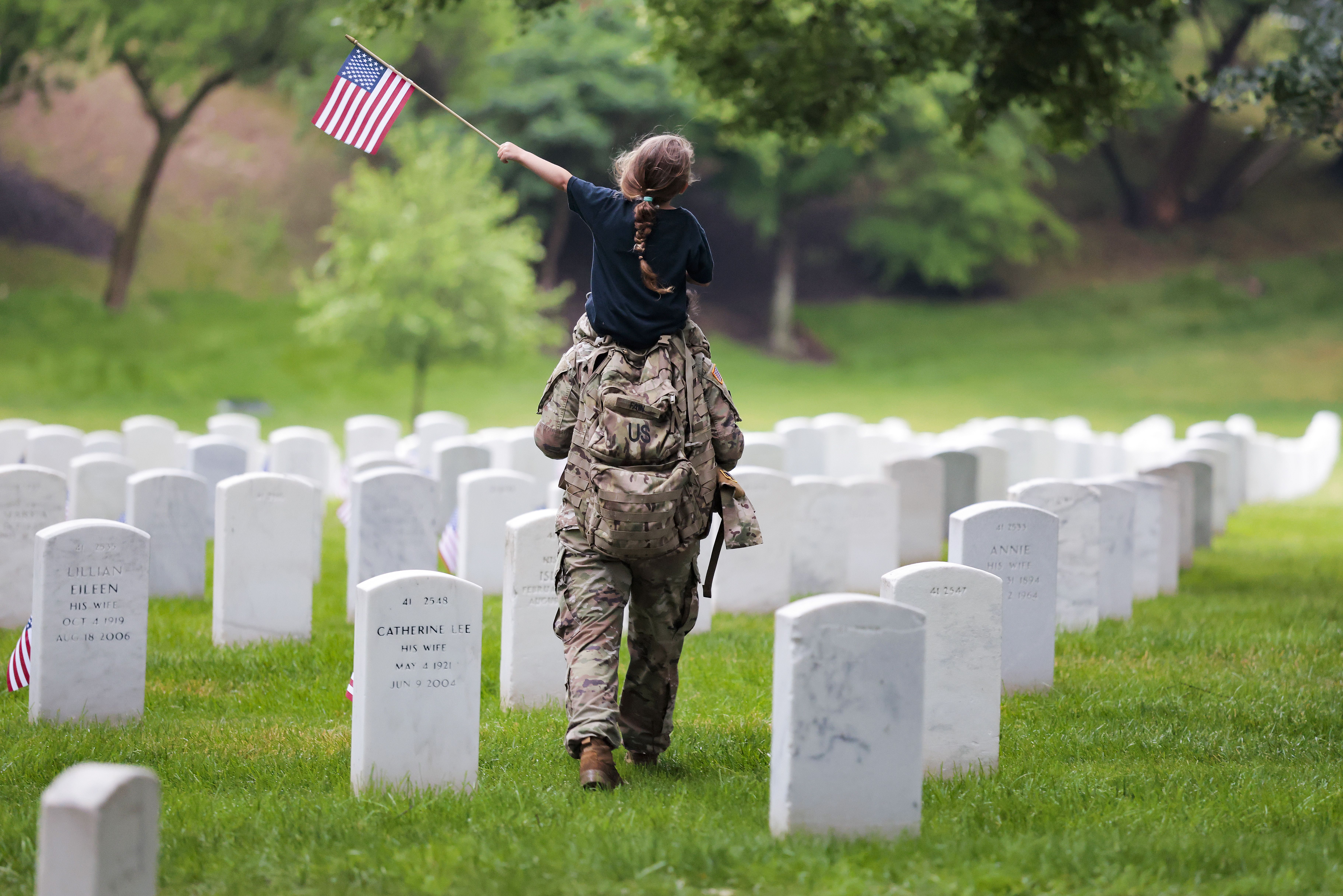 ARLINGTON, VIRGINIA - MAY 21: A member of the 3rd U.S. Infantry Regiment with her daughter on her shoulders places flags in front of each headstone at Arlington National Cemetery on May 21, 2026 in Arlington, Virginia. Before dawn soldiers begin the process of placing a flag in front of approximatel